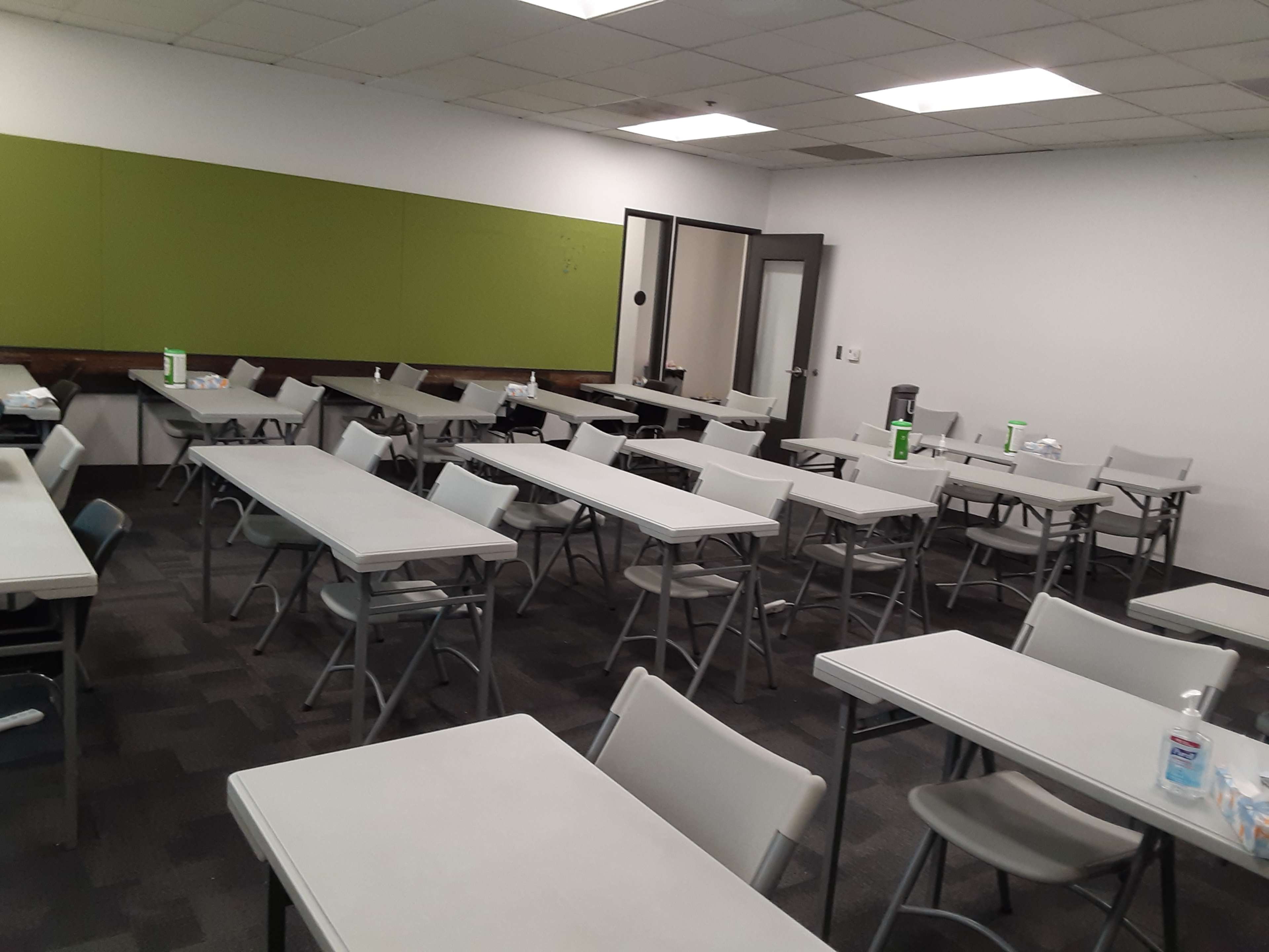 A classroom setting with multiple rows of gray folding tables and chairs, featuring a green bulletin board and supplies on the tables.