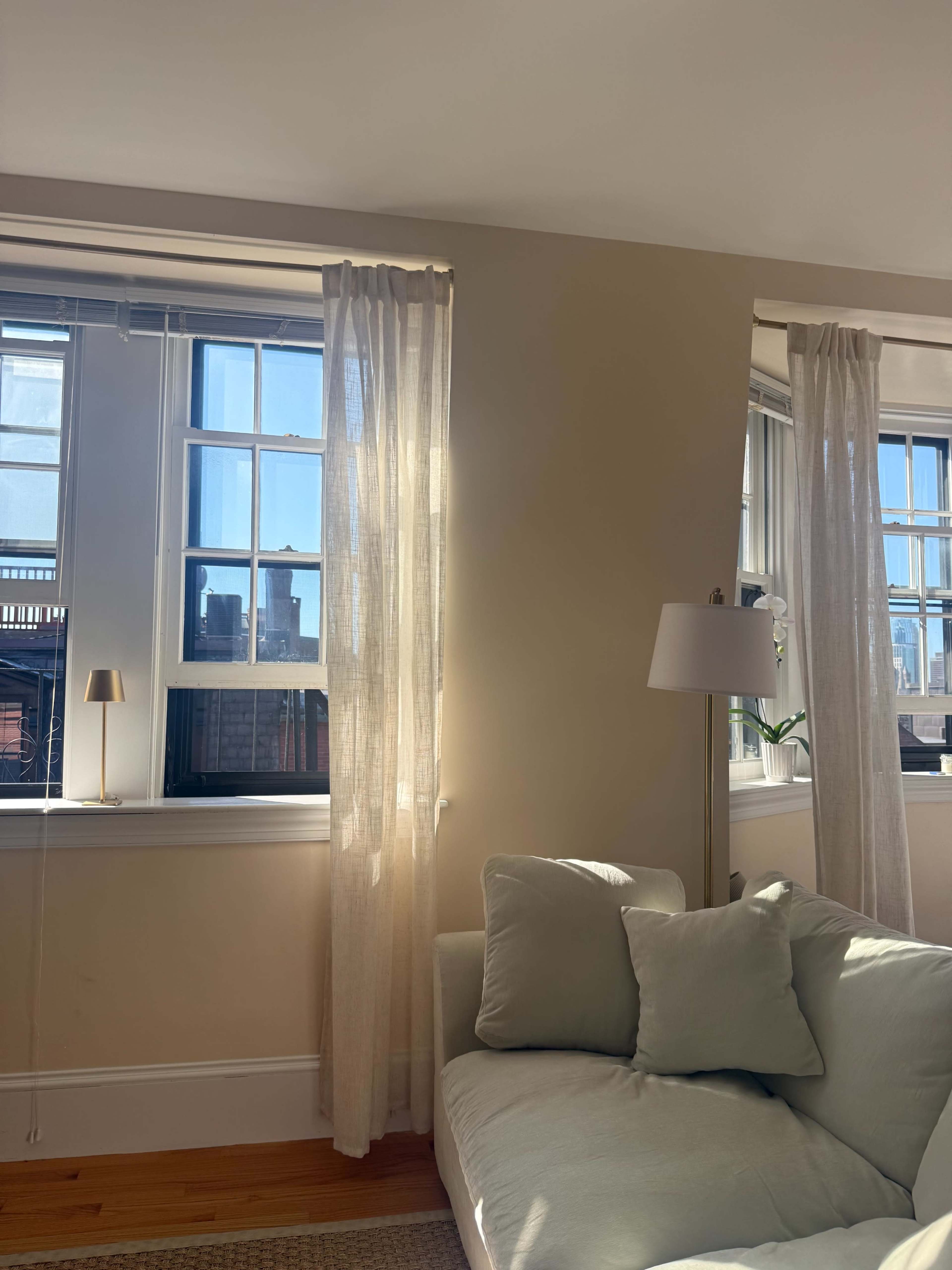 The image shows a well-lit living room with light-colored curtains framing two windows, a small lamp on a side table, and a light green sofa.
