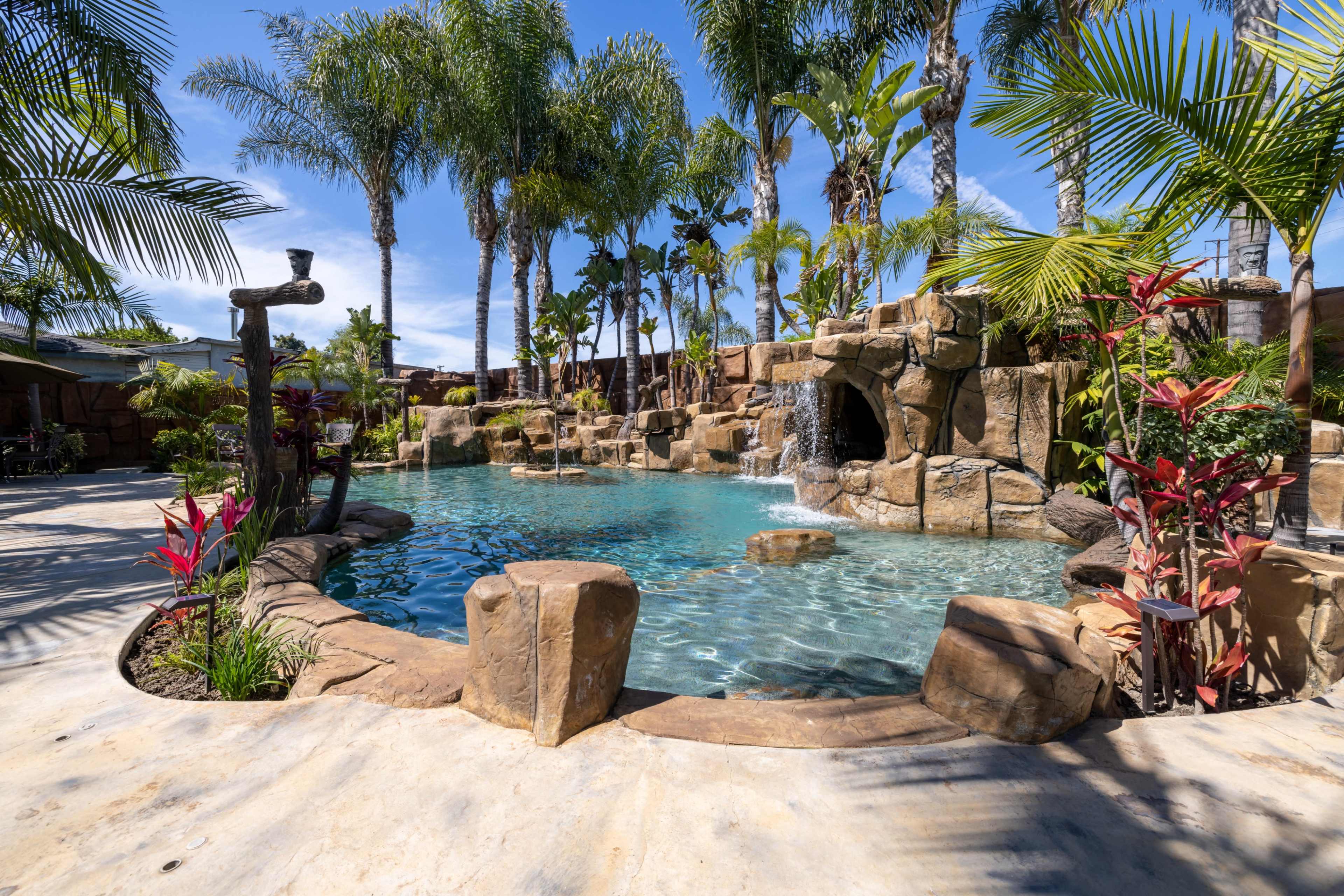 A landscaped pool area surrounded by palm trees and rocks, featuring a small waterfall and vibrant plants.