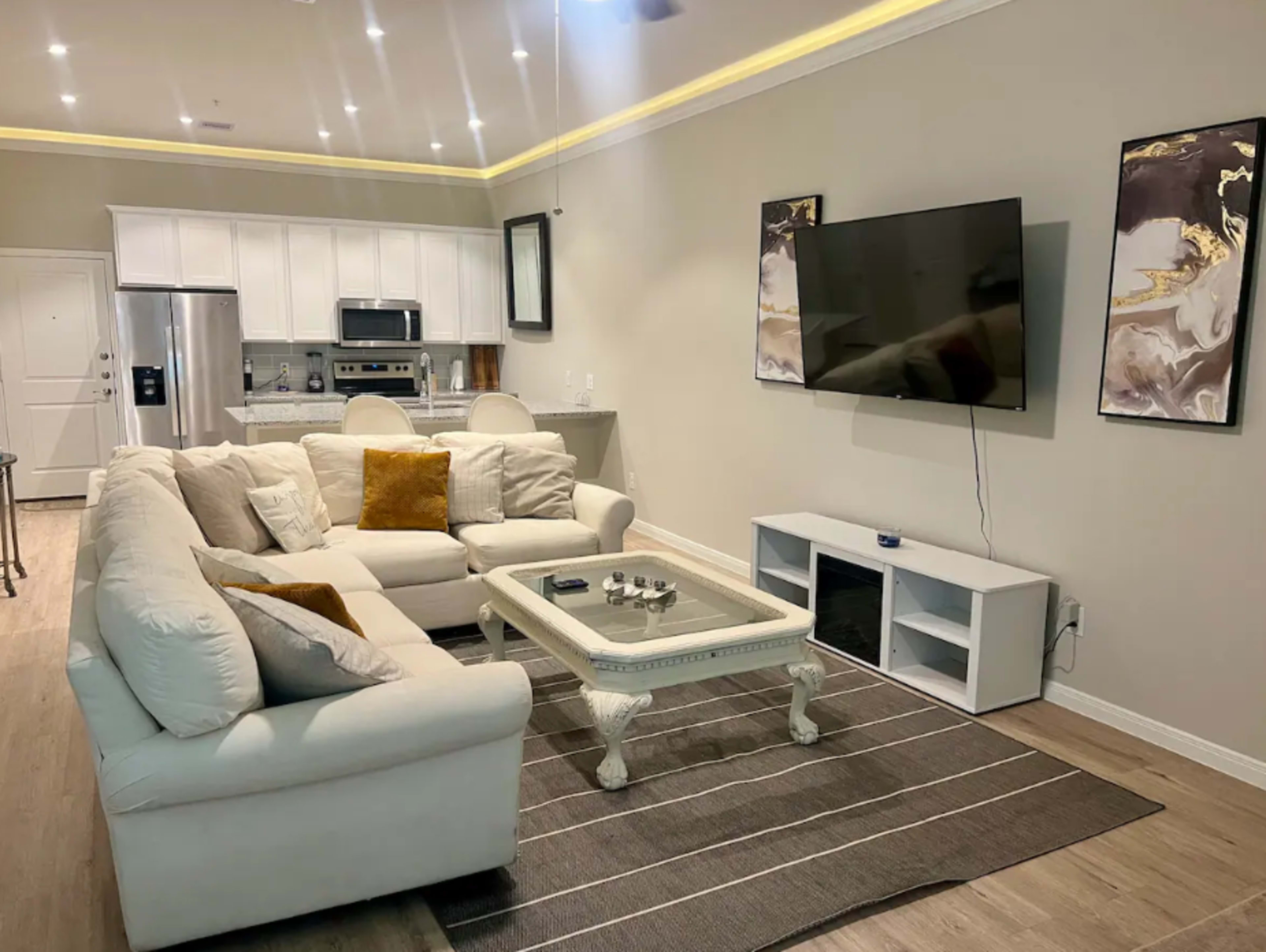 A modern living room featuring a sectional sofa, a coffee table, a television mounted on the wall, and a kitchen area in the background.