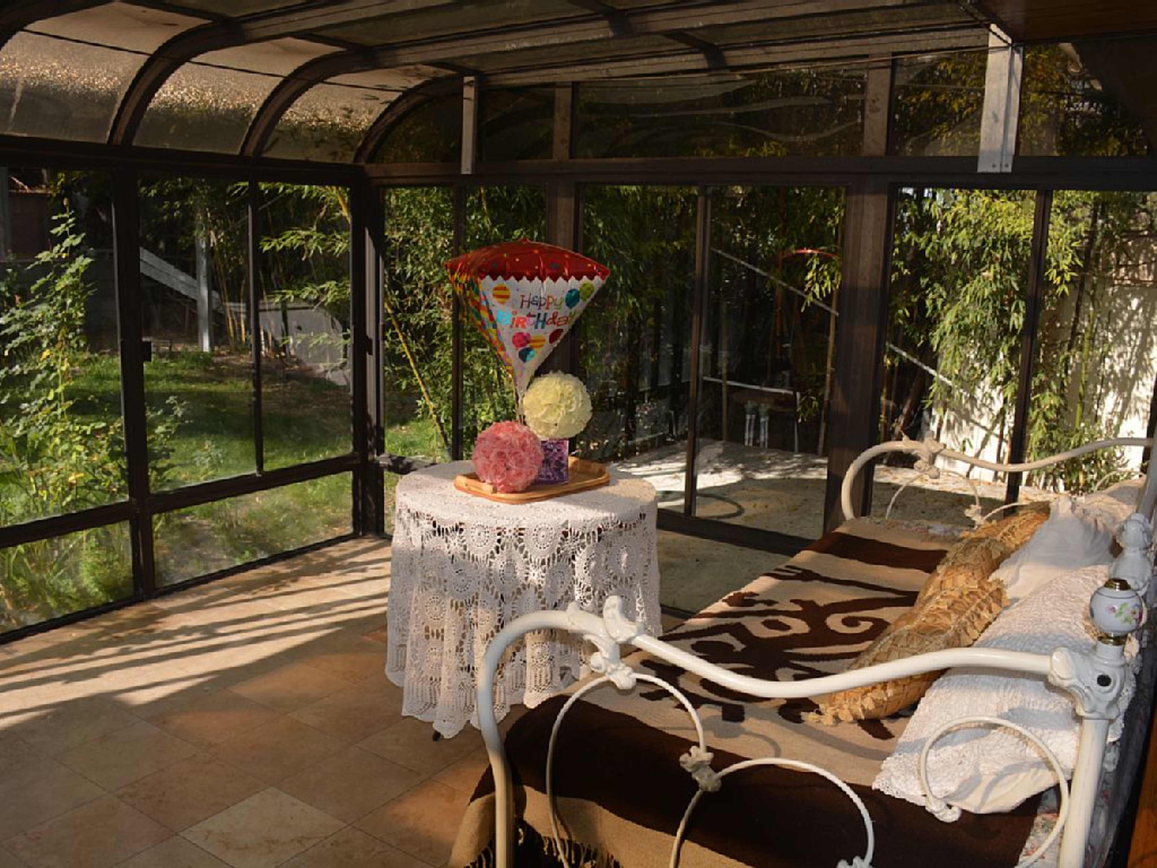 The image shows a sunlit, enclosed porch featuring a table with a decorative tablecloth, a large colorful cone, and two round decorative objects.