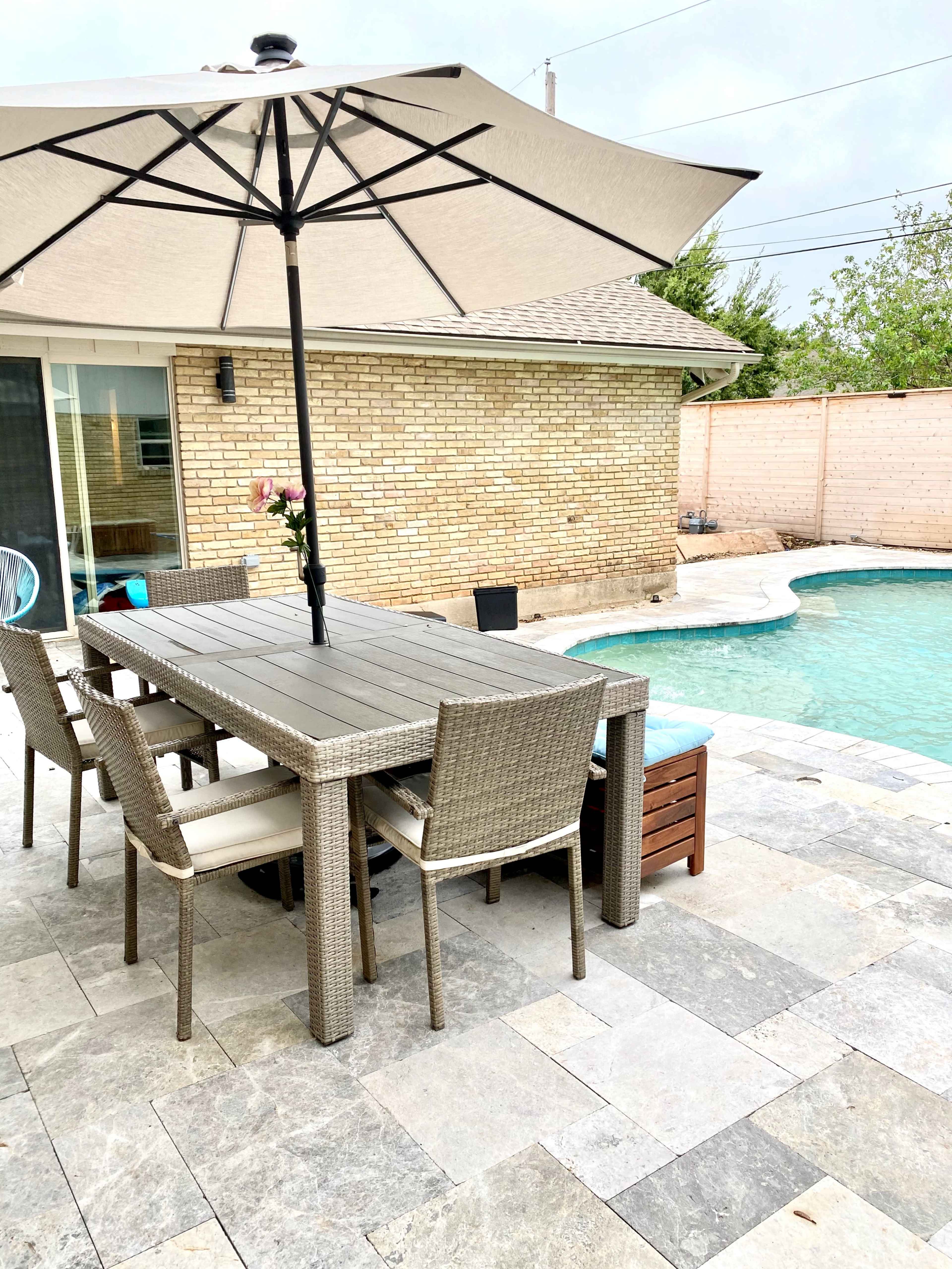 A dining table with chairs is set next to a pool, shaded by a large umbrella, in a backyard with stone paving.