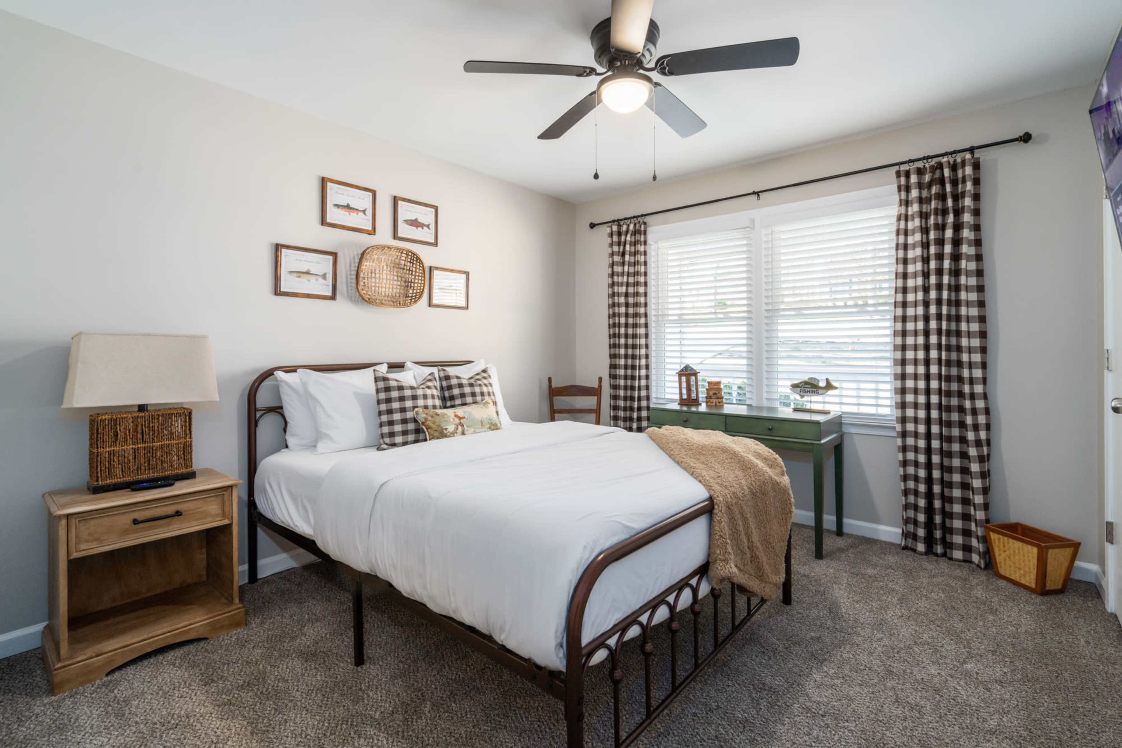 A tidy bedroom featuring a metal bed frame, patterned throw pillows, a nightstand with a lamp, and a green desk by a window with checked curtains.