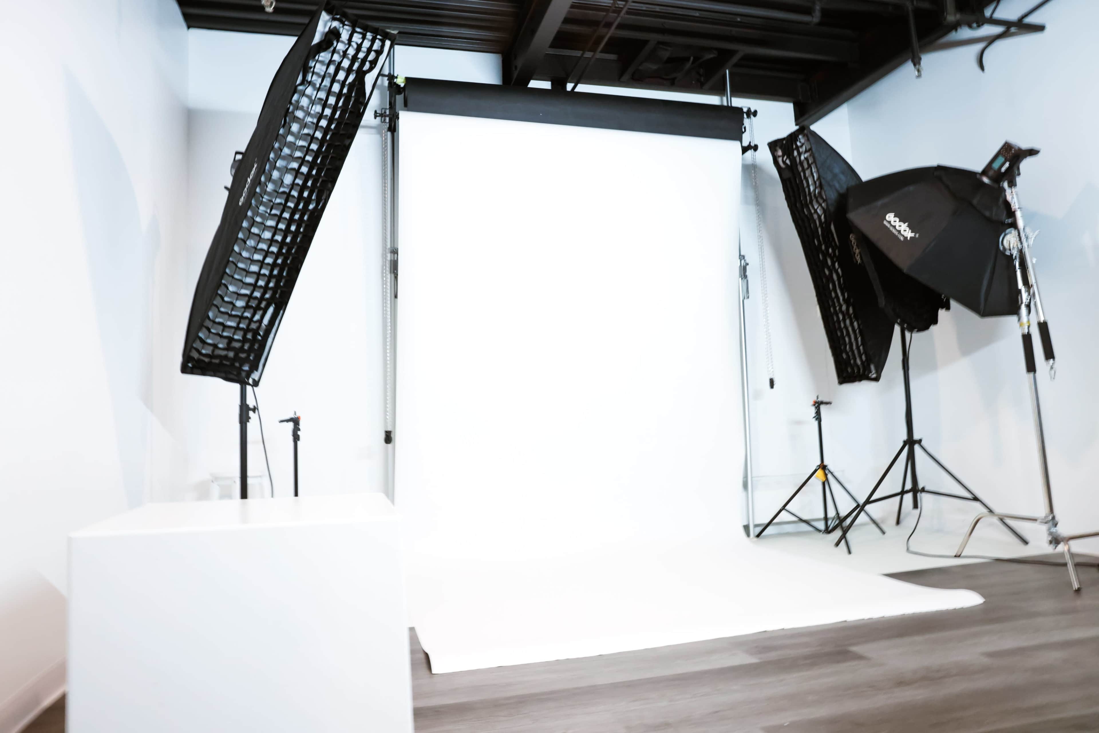 A photography studio features a plain white backdrop with two light sources positioned on either side.