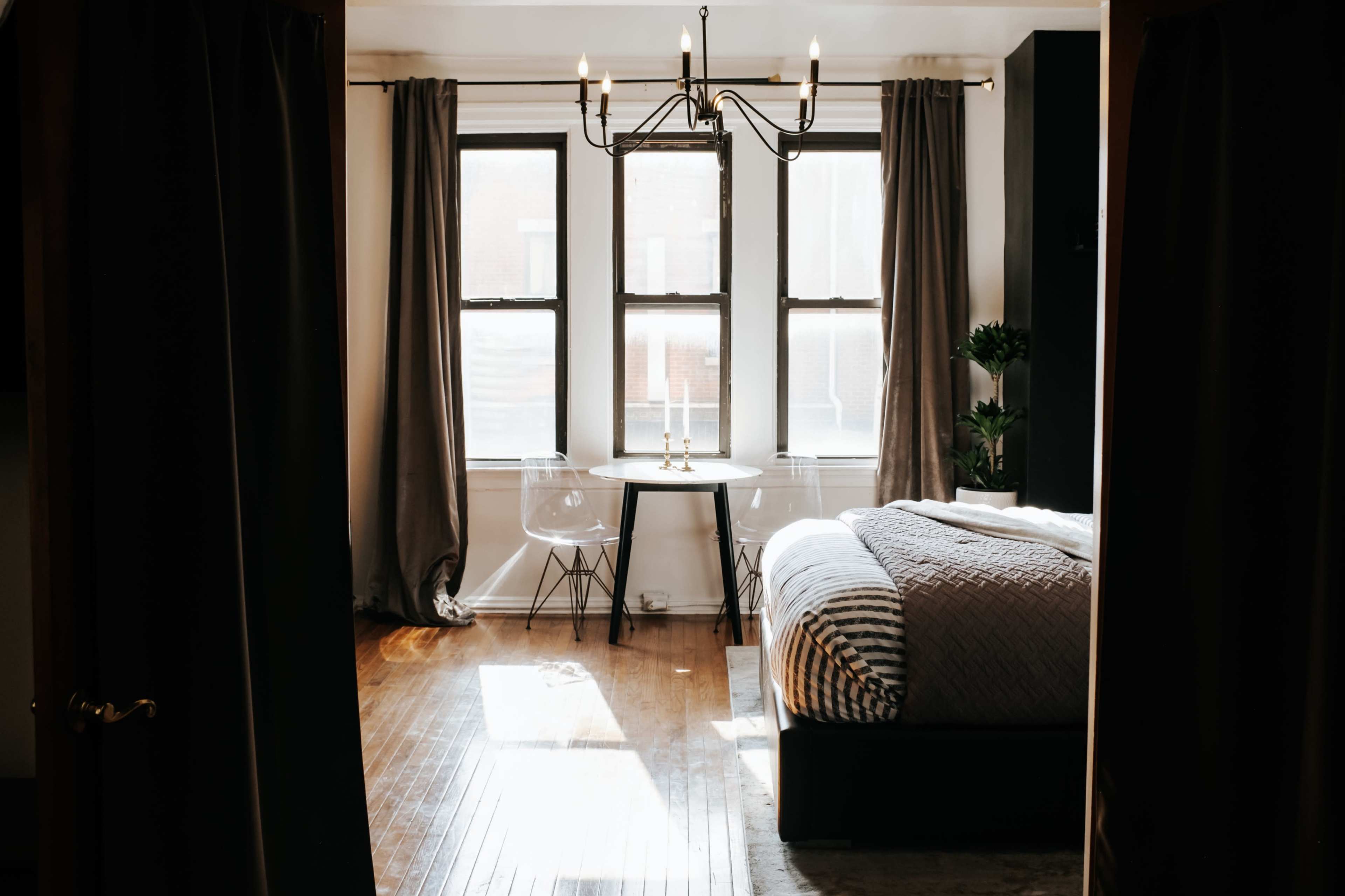 A bedroom features a bed, a small dining table with chairs, and large windows covered with long curtains.