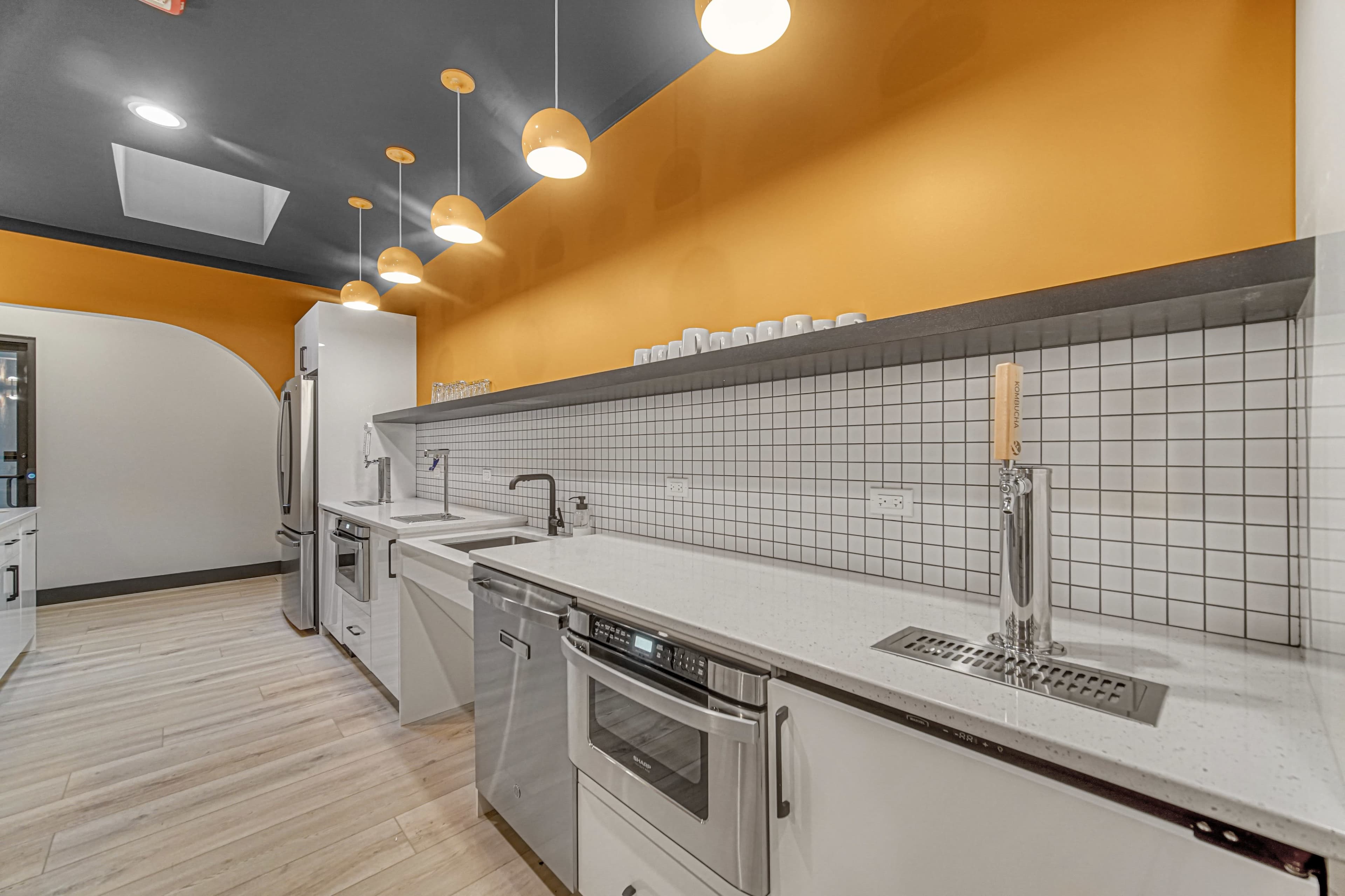 The image shows a modern kitchen with orange walls, white cabinets, and stainless steel appliances, featuring a long countertop with sinks and a wall-mounted beer tap.