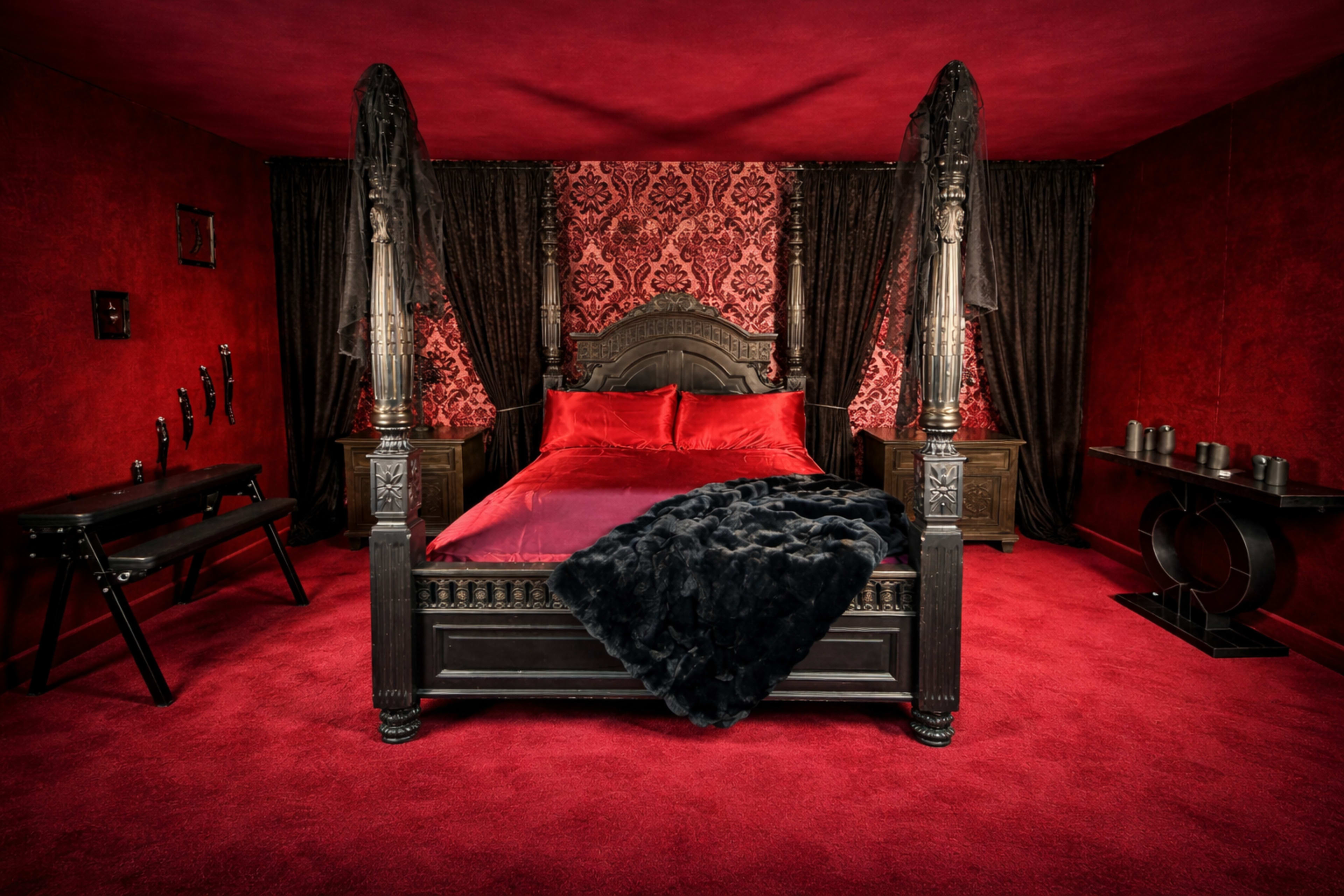 The image depicts a bedroom with red walls and carpet, featuring a black four-poster bed with a red bedding set and dark curtains.