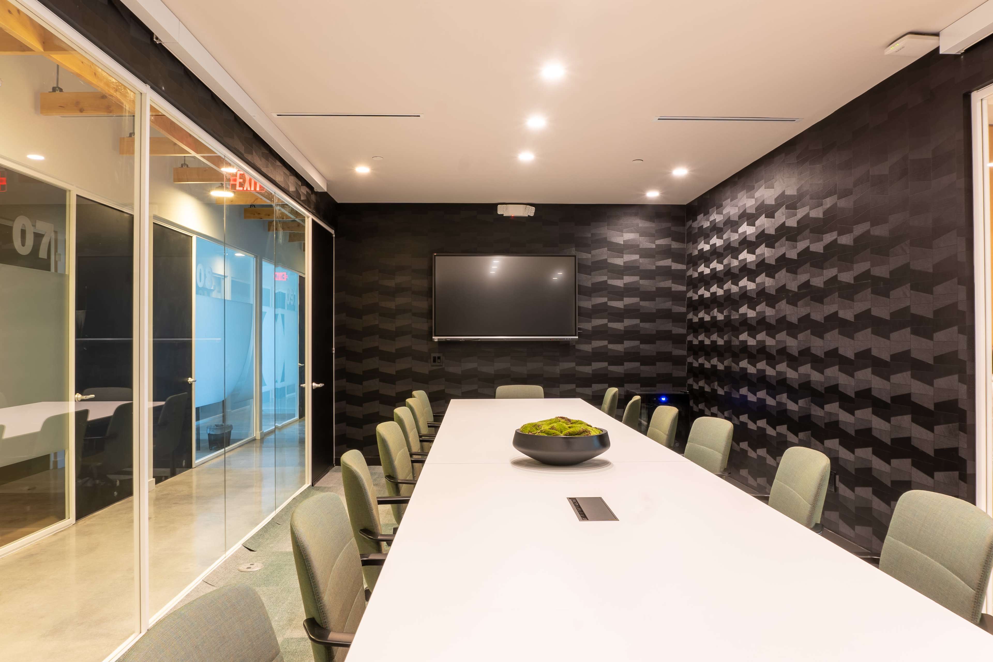 A modern conference room features a long white table surrounded by green chairs, with a large screen mounted on a textured black wall.