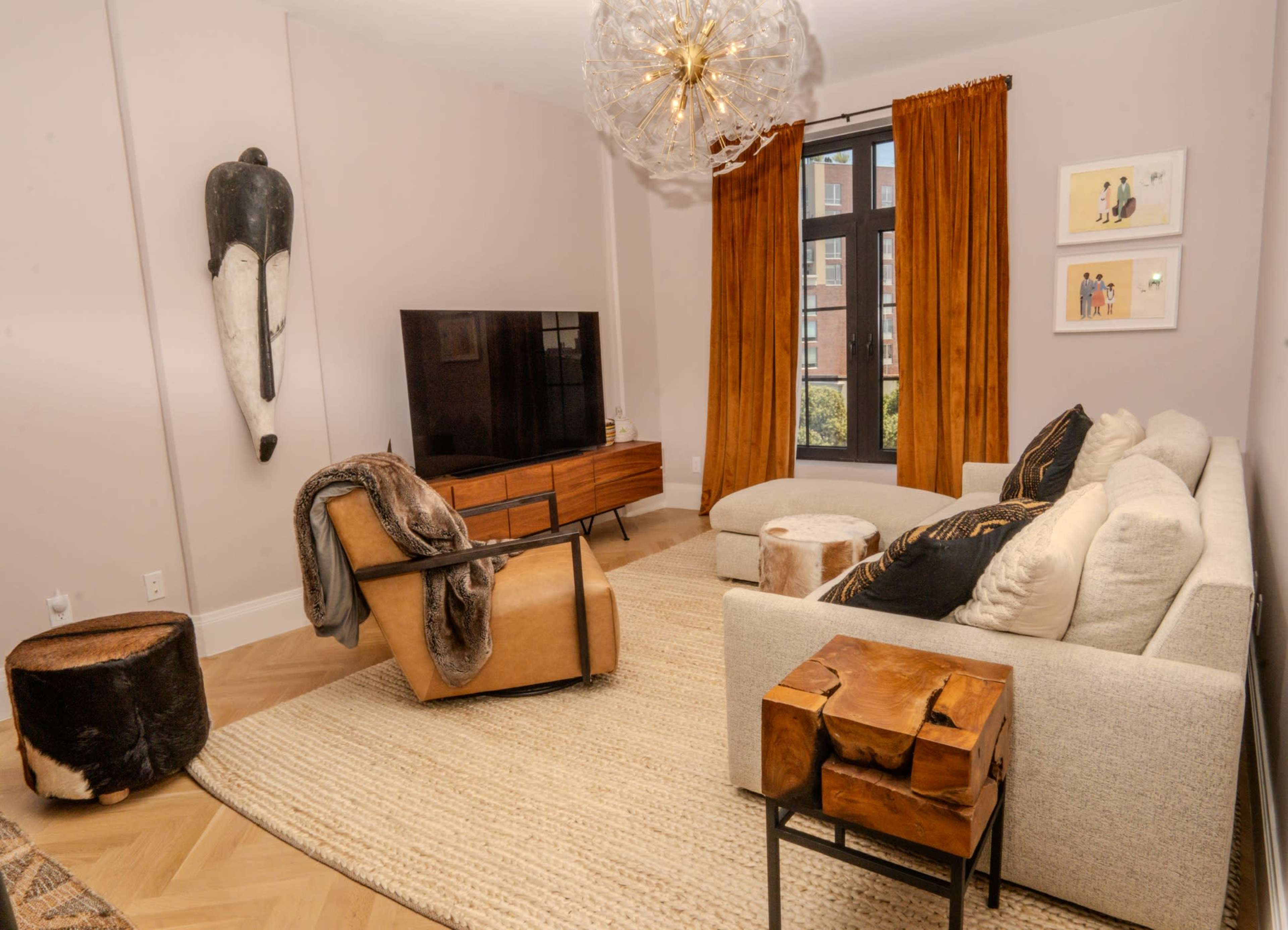 A modern living room features a light-colored sofa, a wooden media console with a television, an armchair, and decorative elements including a wall hanging and curtains.