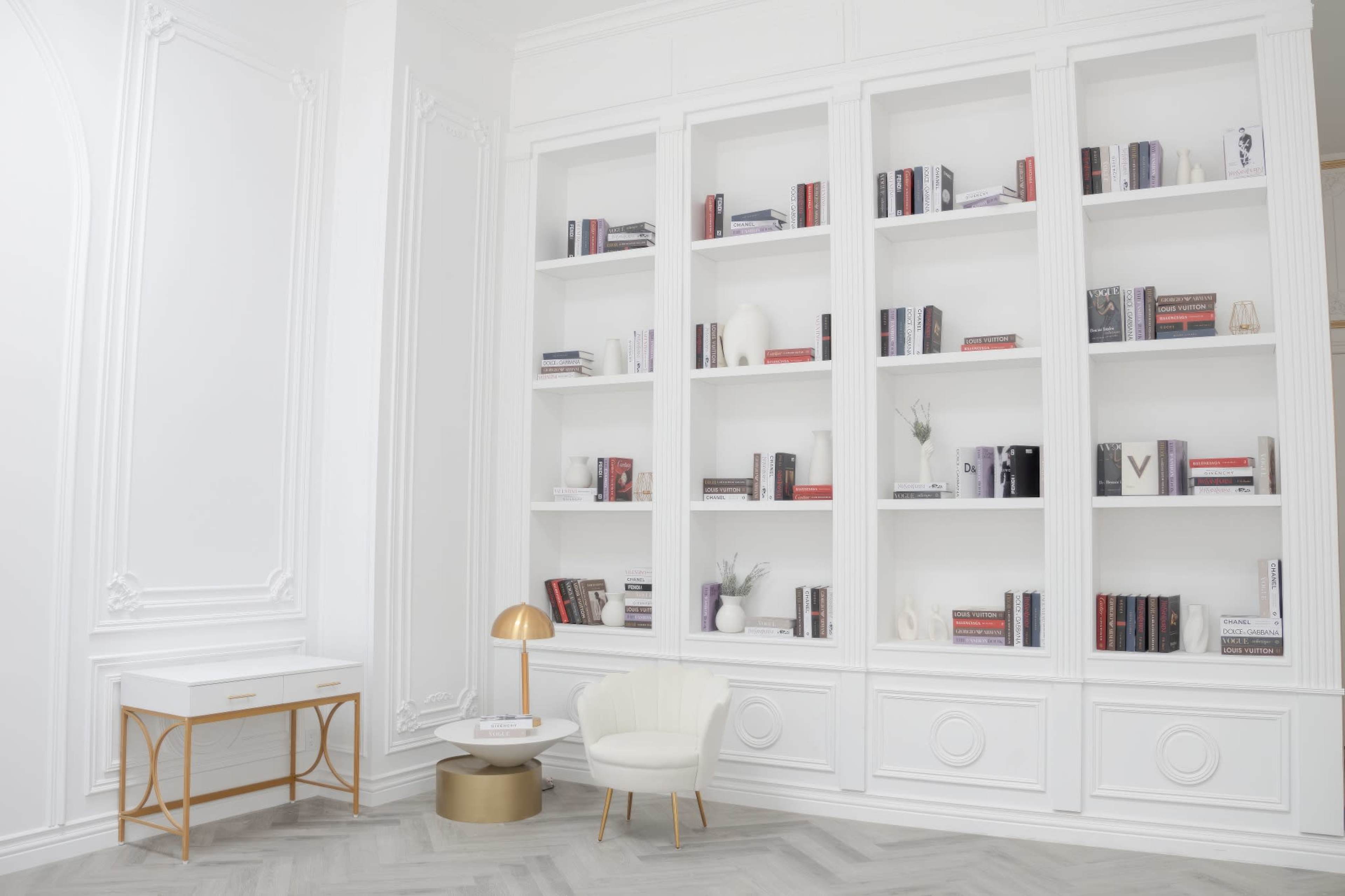 A modern interior space featuring a white bookshelf filled with books, a small round table, a chair, and a desk with a lamp against a light-colored wall.
