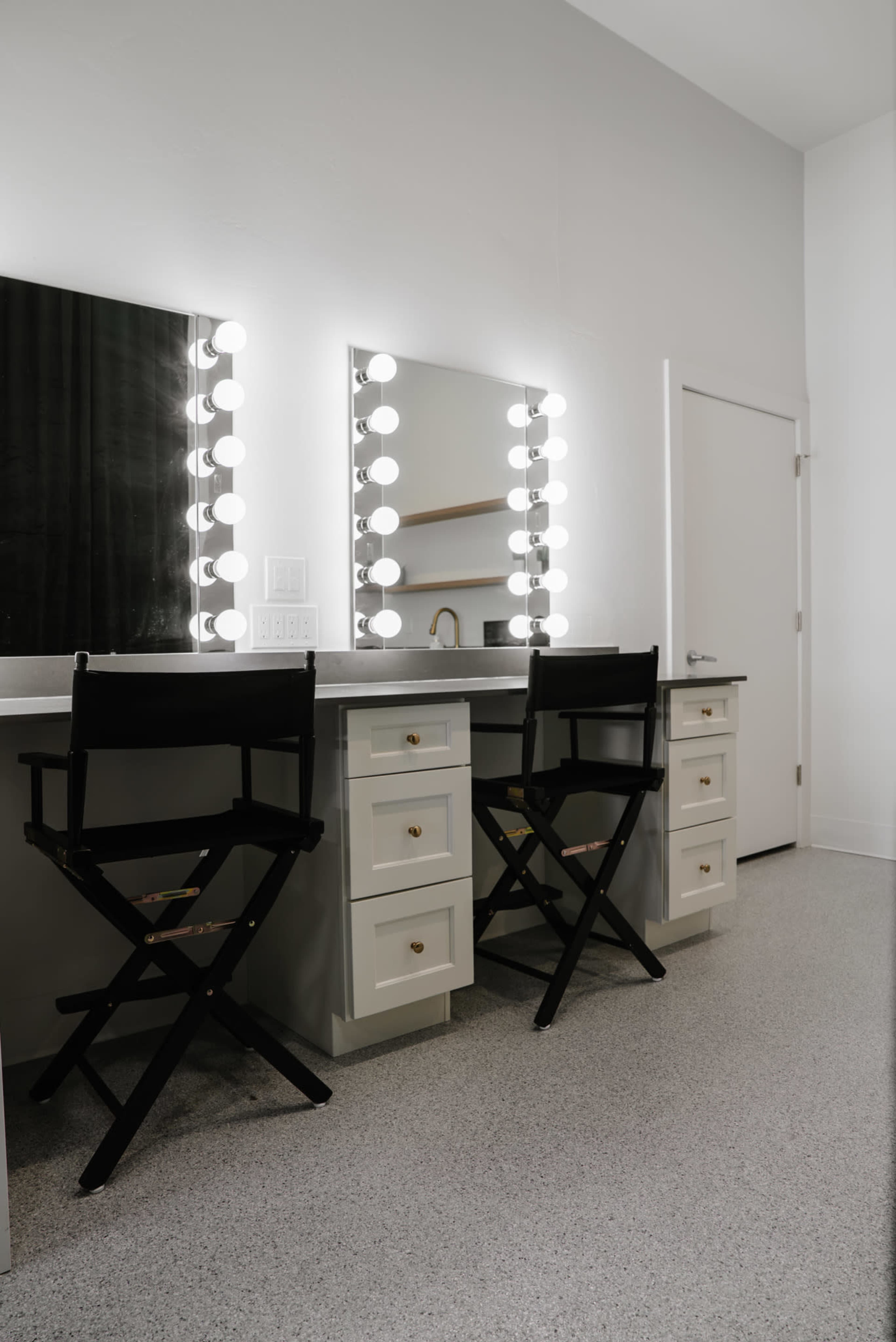 The image shows a well-lit vanity area with two black director's chairs, a mirror with round light bulbs, and a white countertop featuring drawers.