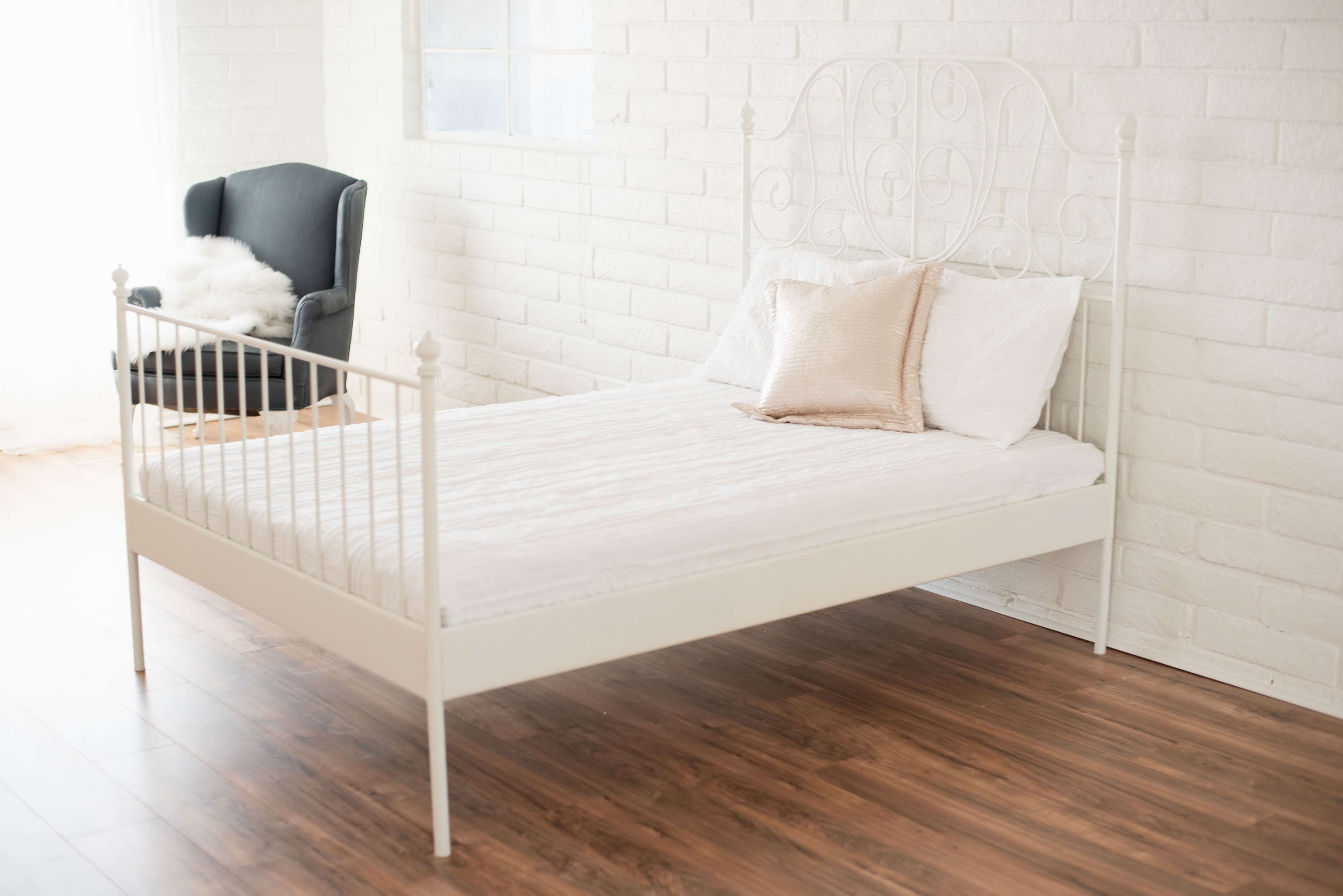 A white metal bed with a simple headboard sits on a wooden floor near a black armchair in a bright, minimalist room.