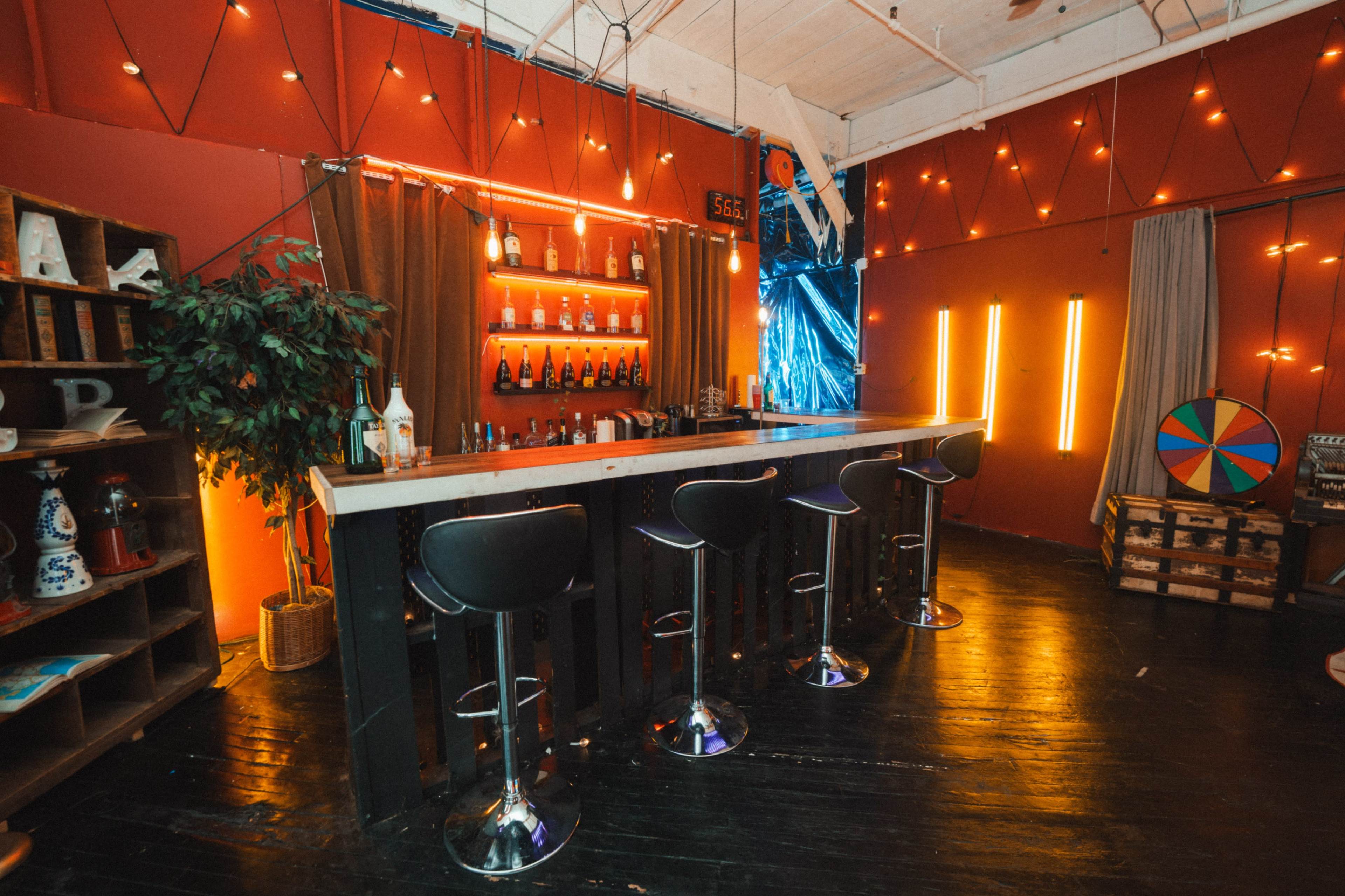 The image shows a modern bar area with several high stools, a wooden counter, shelves of bottles, and decorative lighting against a vibrant red wall.