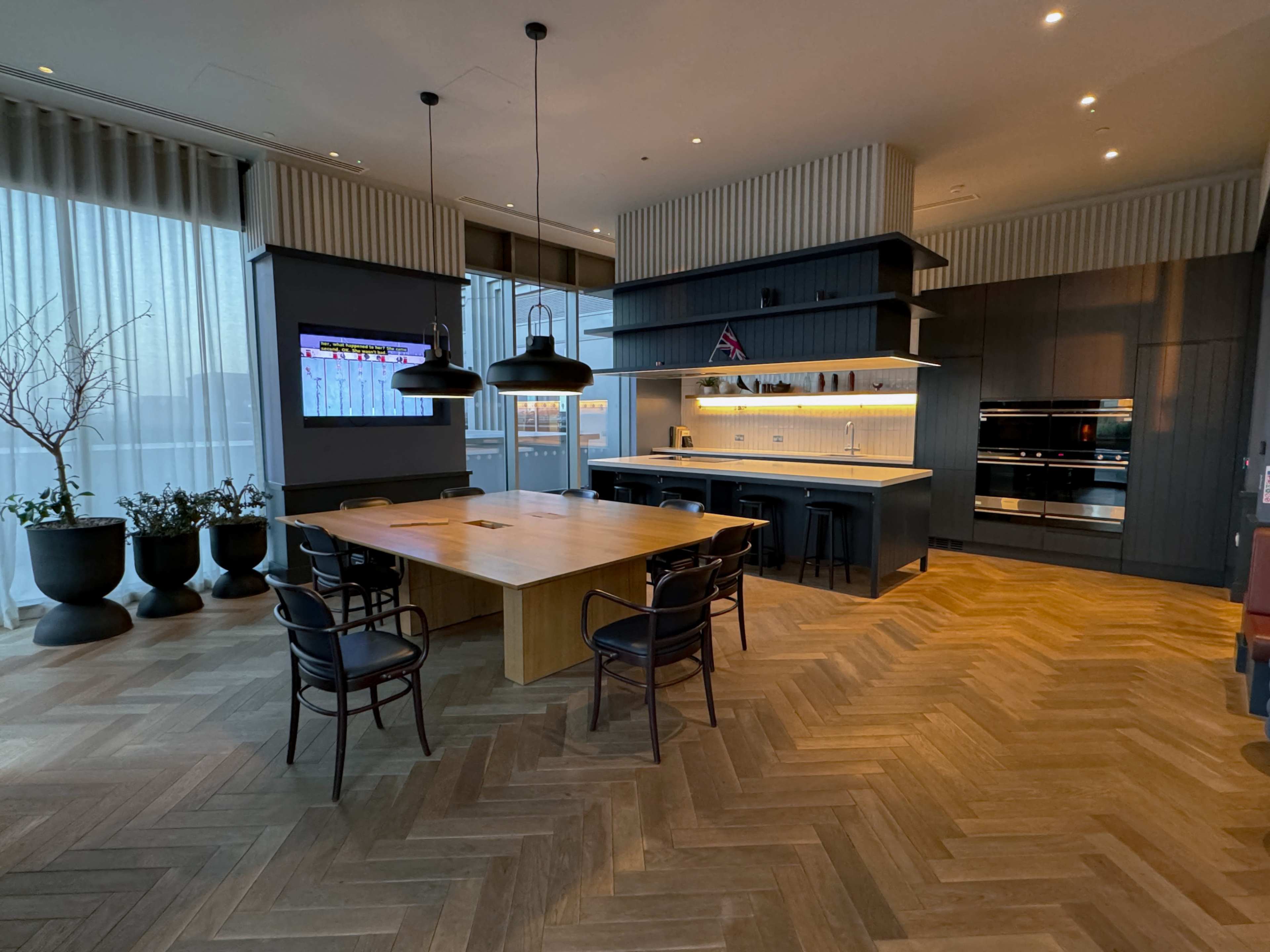 A modern kitchen and dining area with a large wooden table, black chairs, and a wall-mounted TV, illuminated by pendant lights.