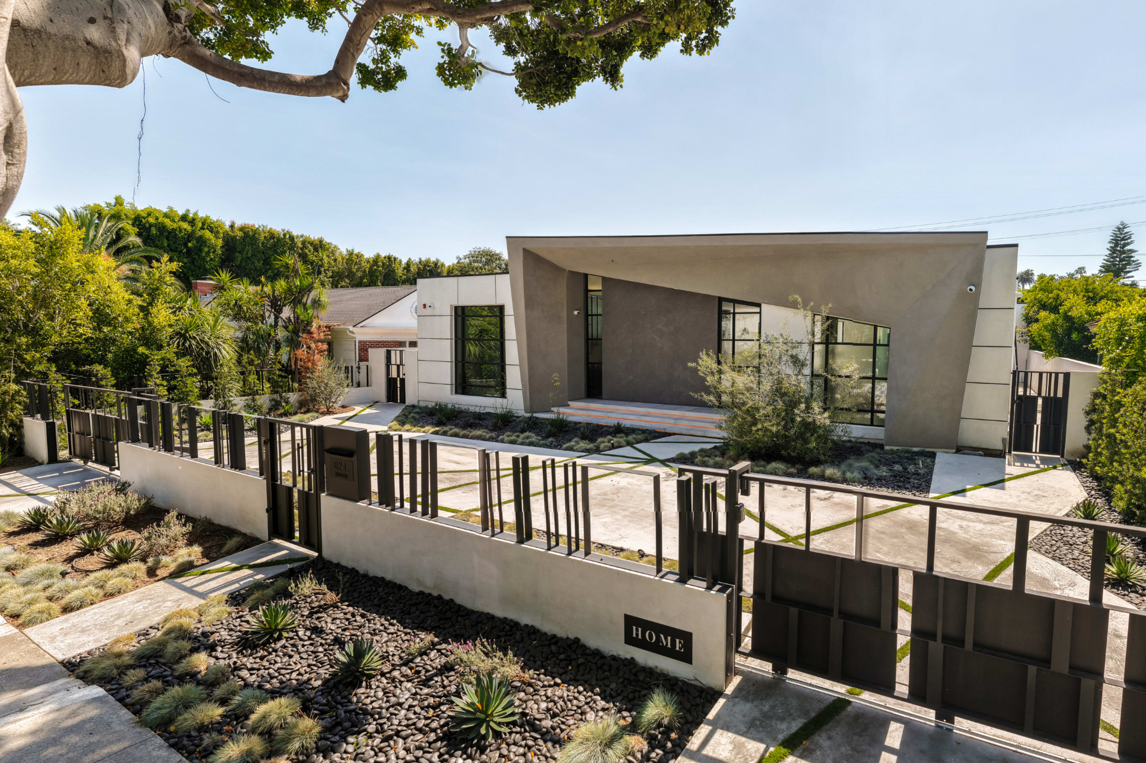 The image shows a contemporary house with a unique architectural design, surrounded by a modern fence and landscaped with various plants.