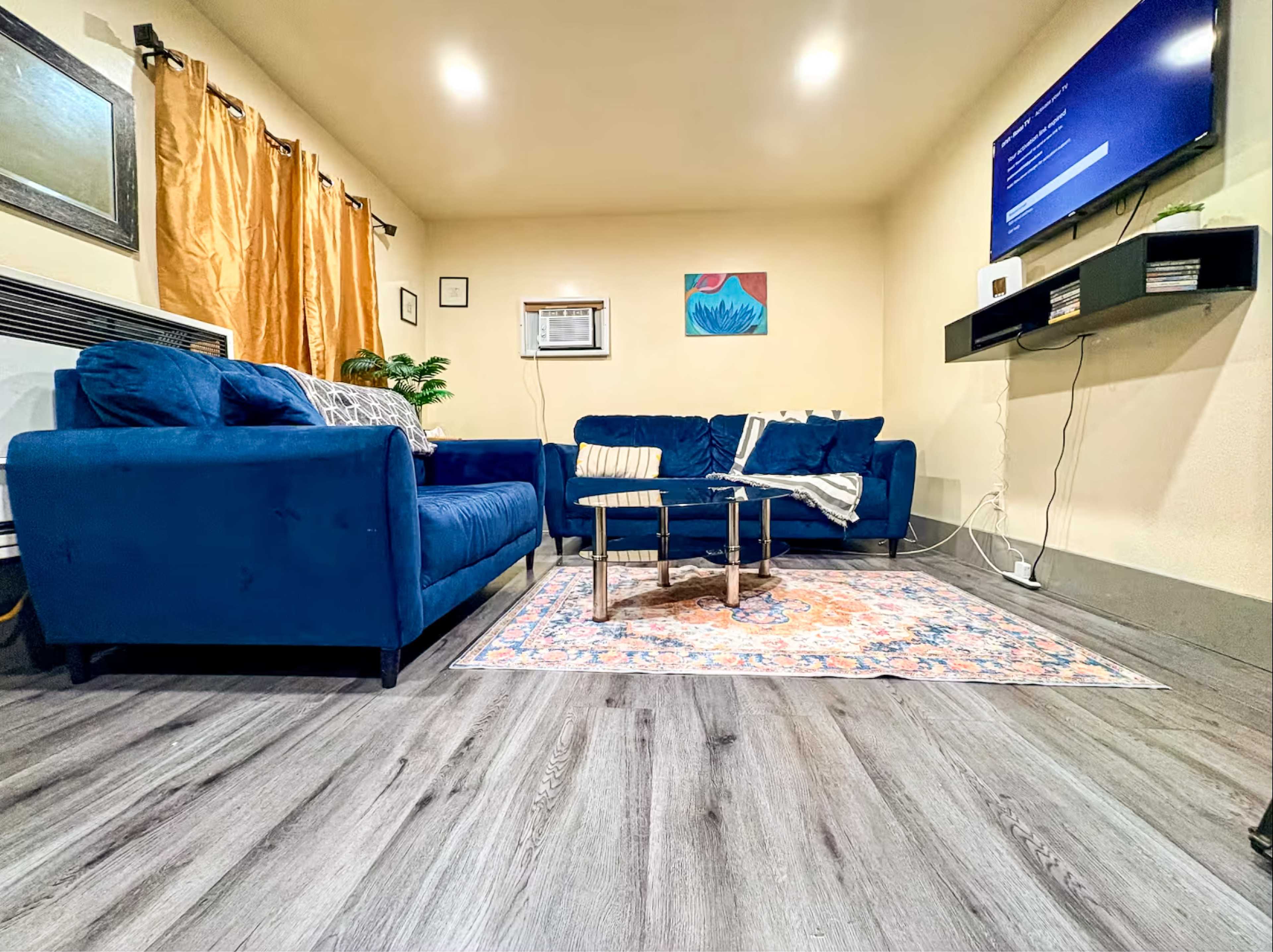 The image shows a cozy living room with blue sofas, a coffee table, a patterned rug, and a wall-mounted TV.