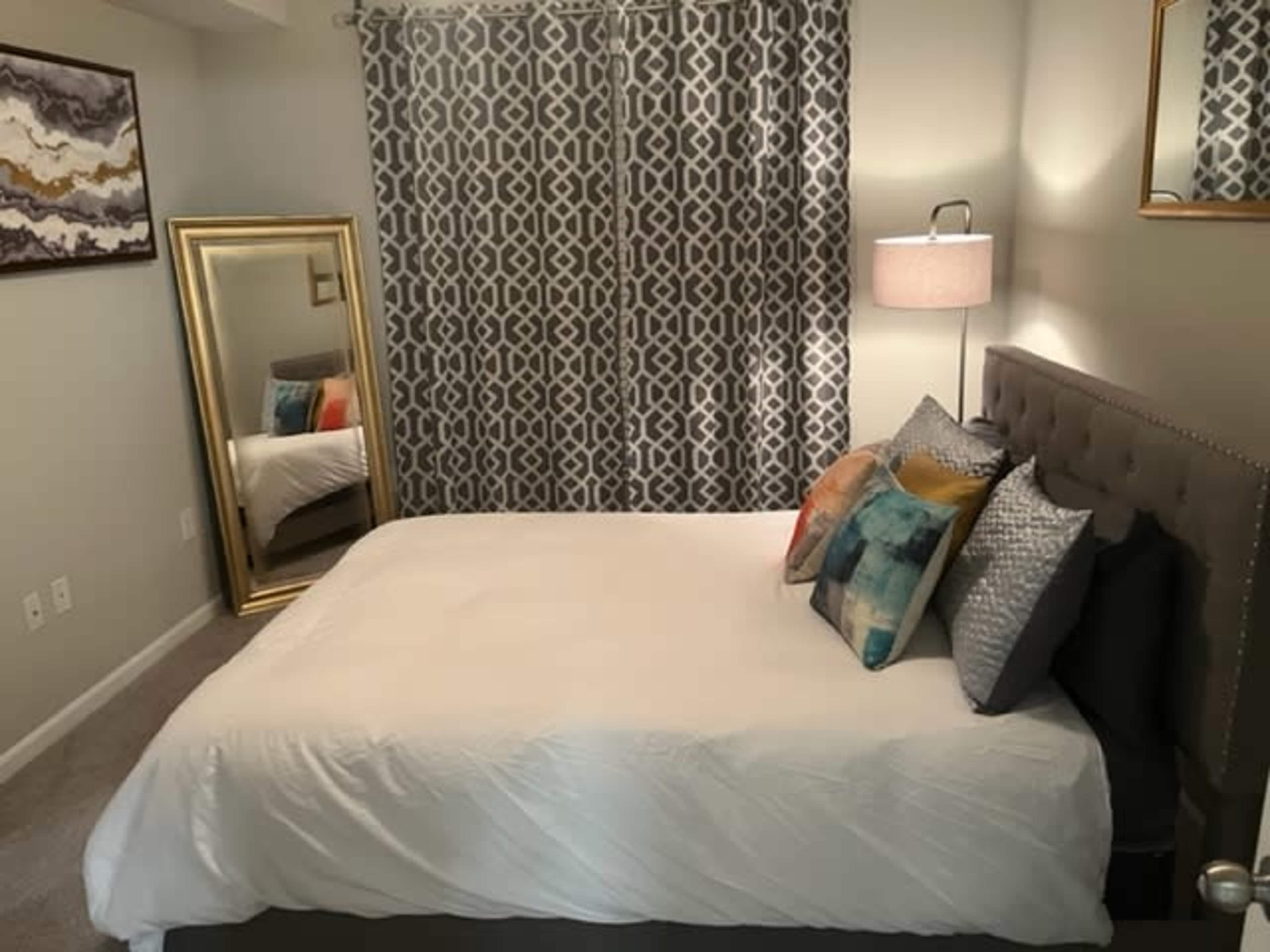 A neatly arranged bedroom features a bed with decorative pillows, a mirrored dresser, and patterned curtains.