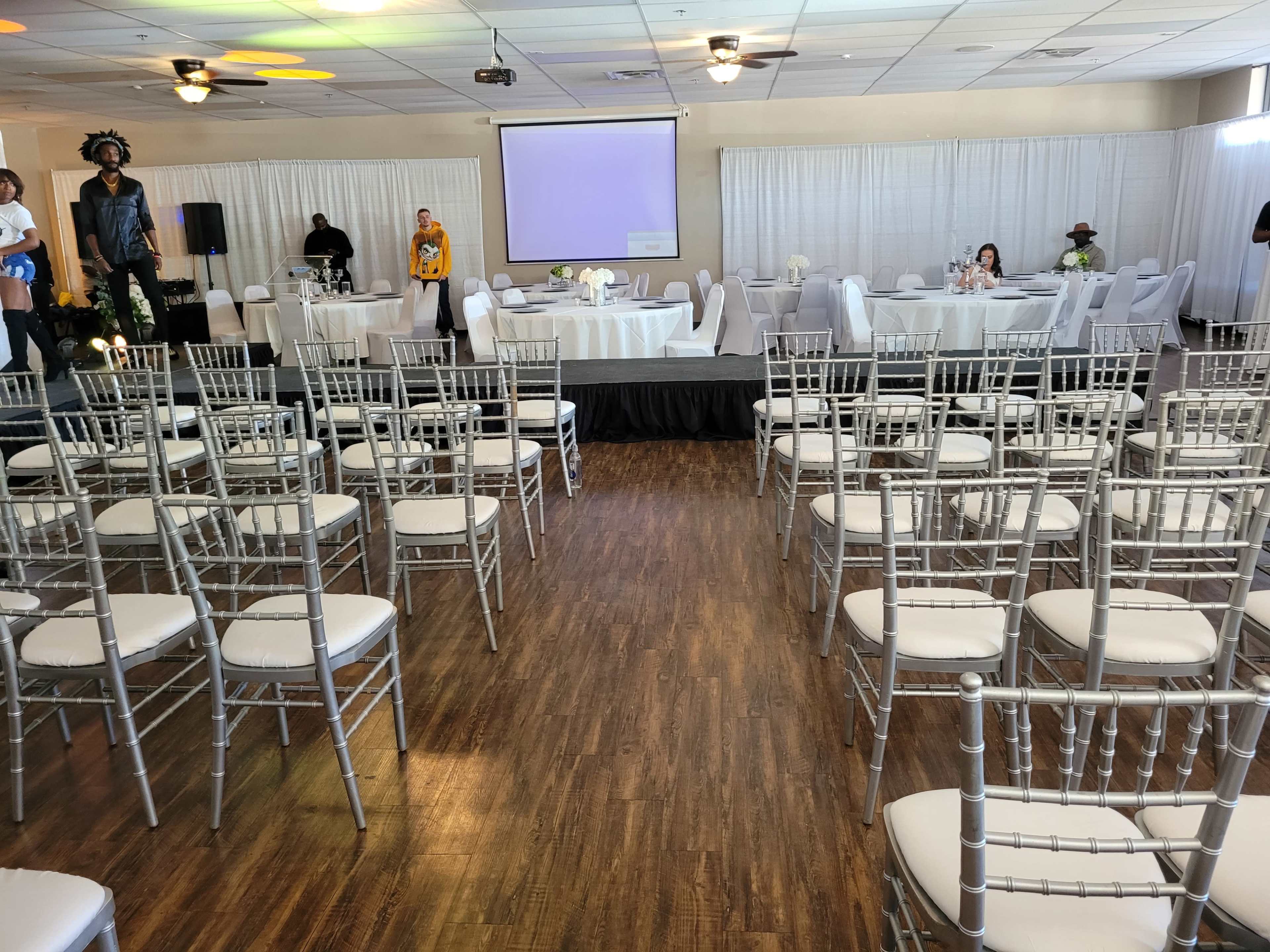 A spacious event hall features neatly arranged silver chiavari chairs facing a small stage, with tables set for dining in the background.