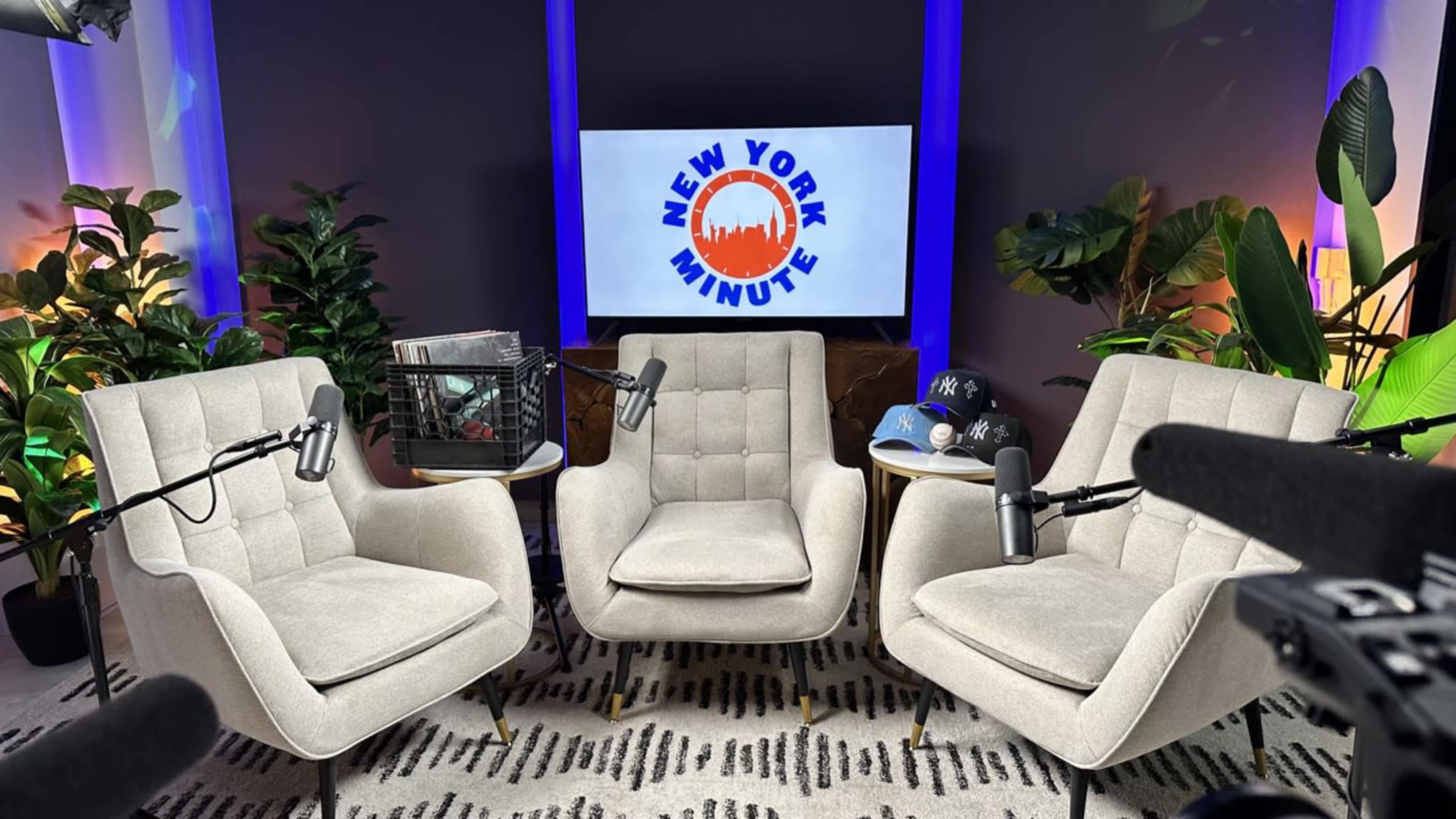 The image shows a modern studio setup with three upholstered chairs facing a screen displaying the "New York Minute" logo, surrounded by potted plants.