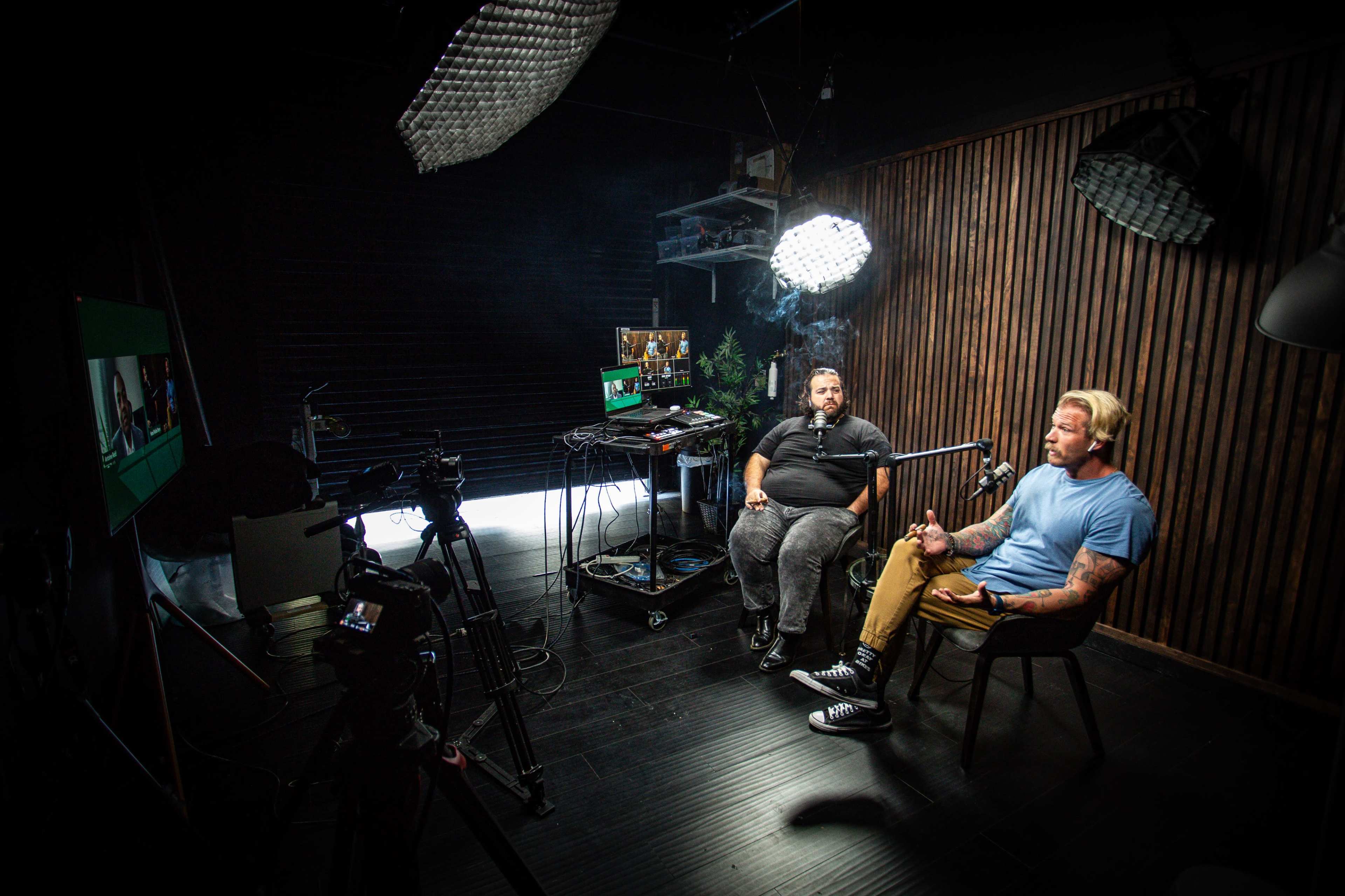 Two men are seated in a dimly lit studio, engaged in a conversation while being recorded by cameras and illuminated by softbox lights.