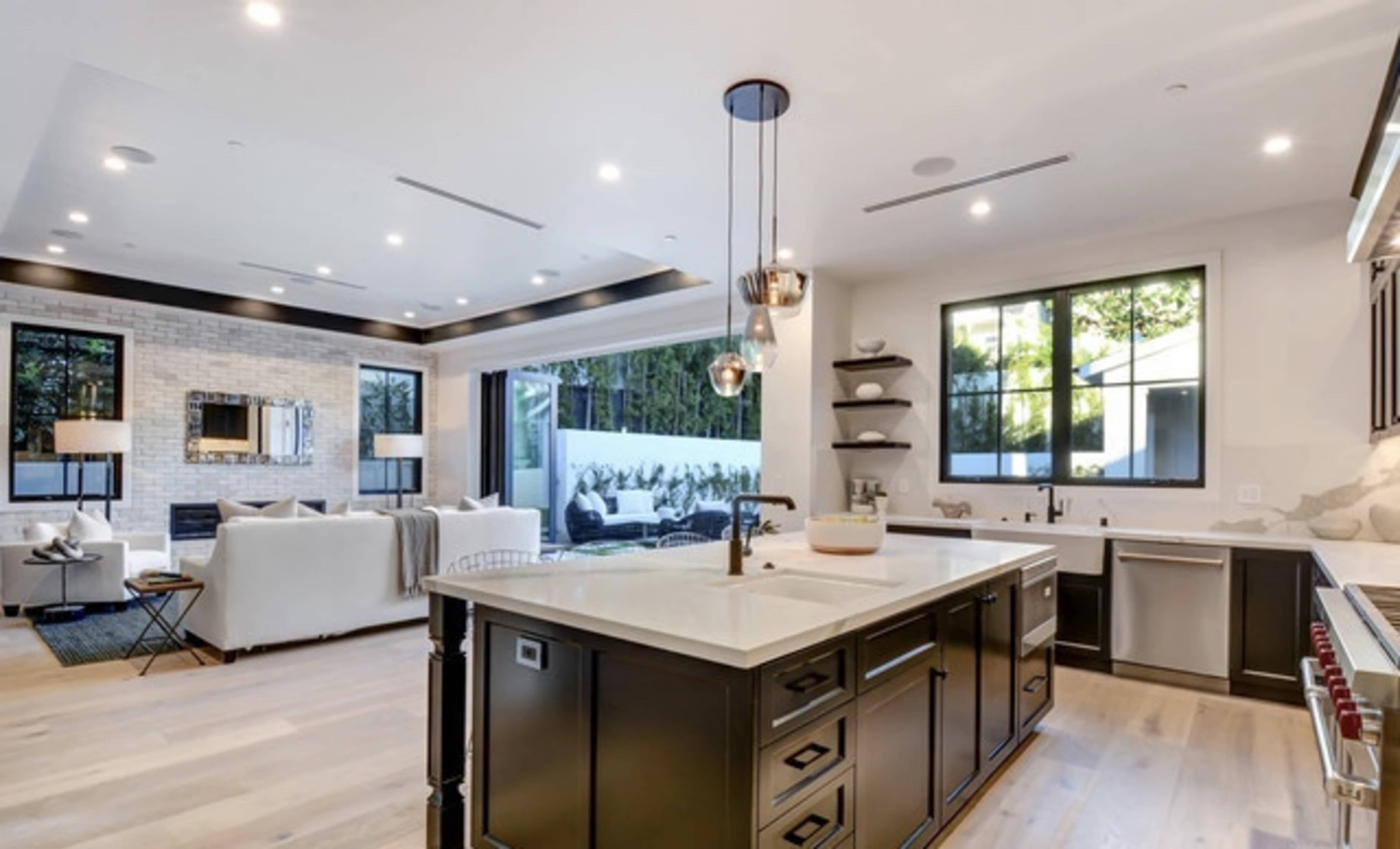 A modern kitchen with an island, stainless steel appliances, and a view of an outdoor seating area through large windows.