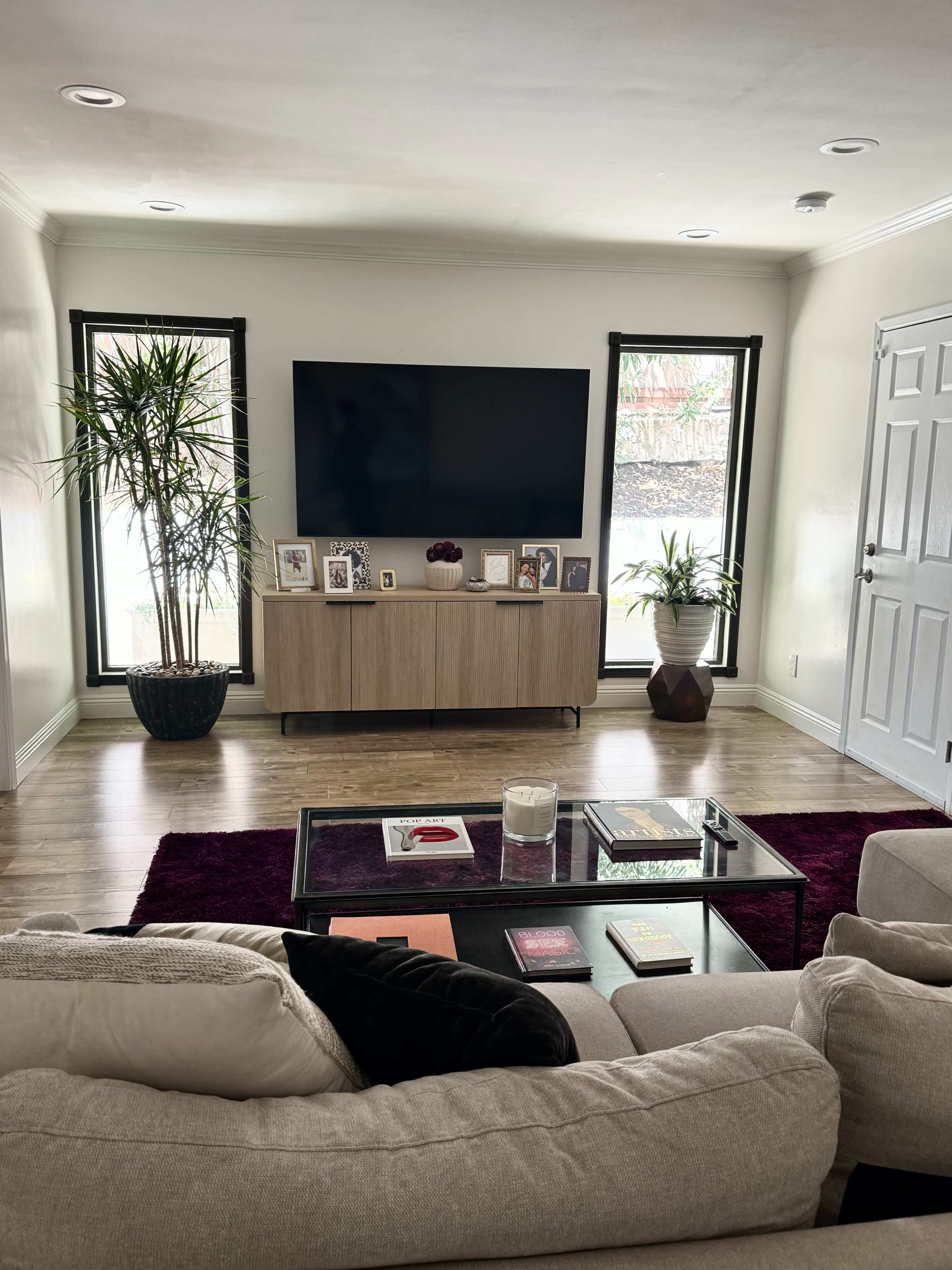 A cozy living room features a large flat-screen TV mounted on a light-colored wall, flanked by windows, with a wooden media console beneath and a coffee table in front of a sectional sofa.