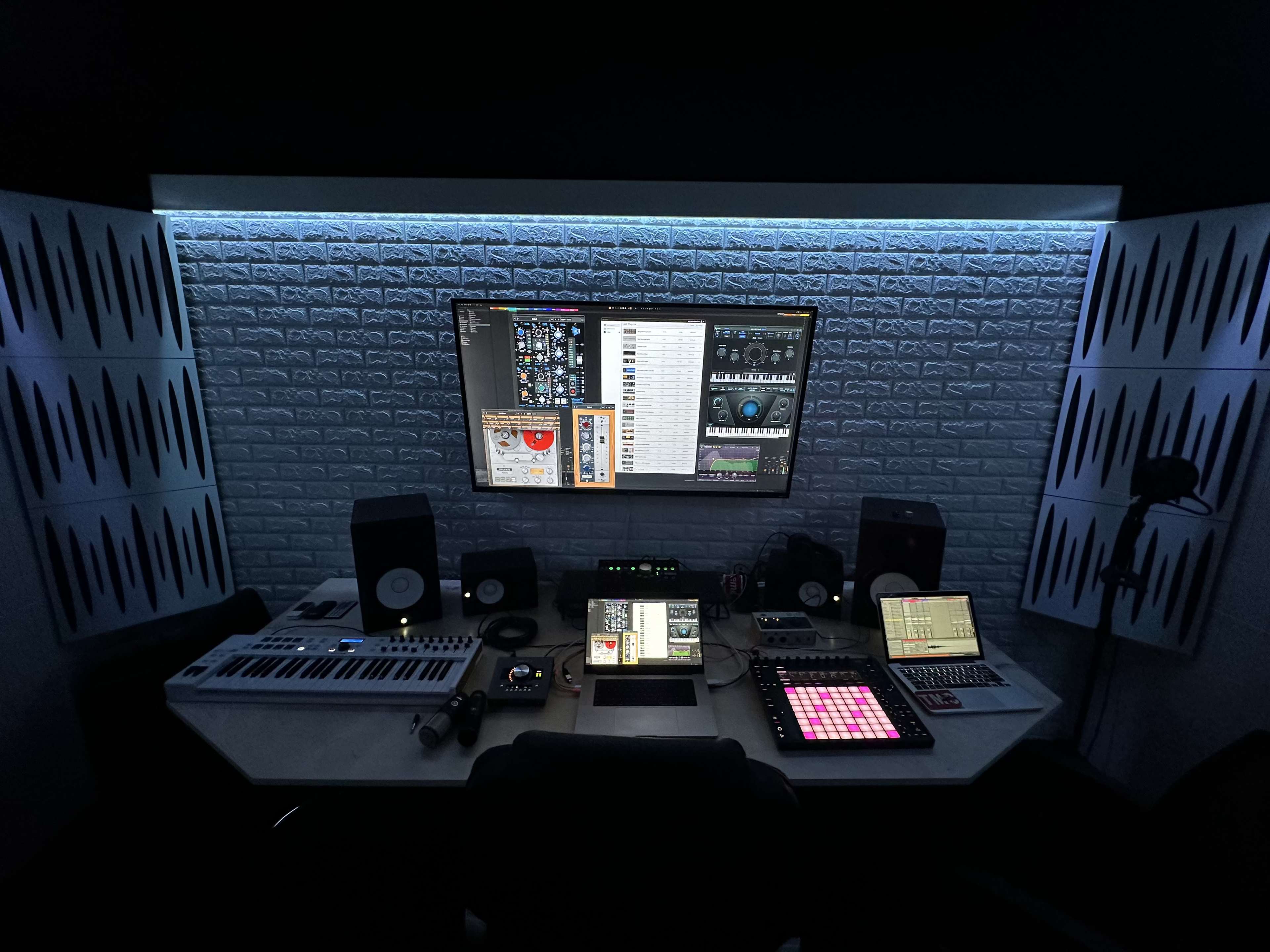 The image shows a modern music production studio with various audio equipment, multiple screens, and a backlit wall.