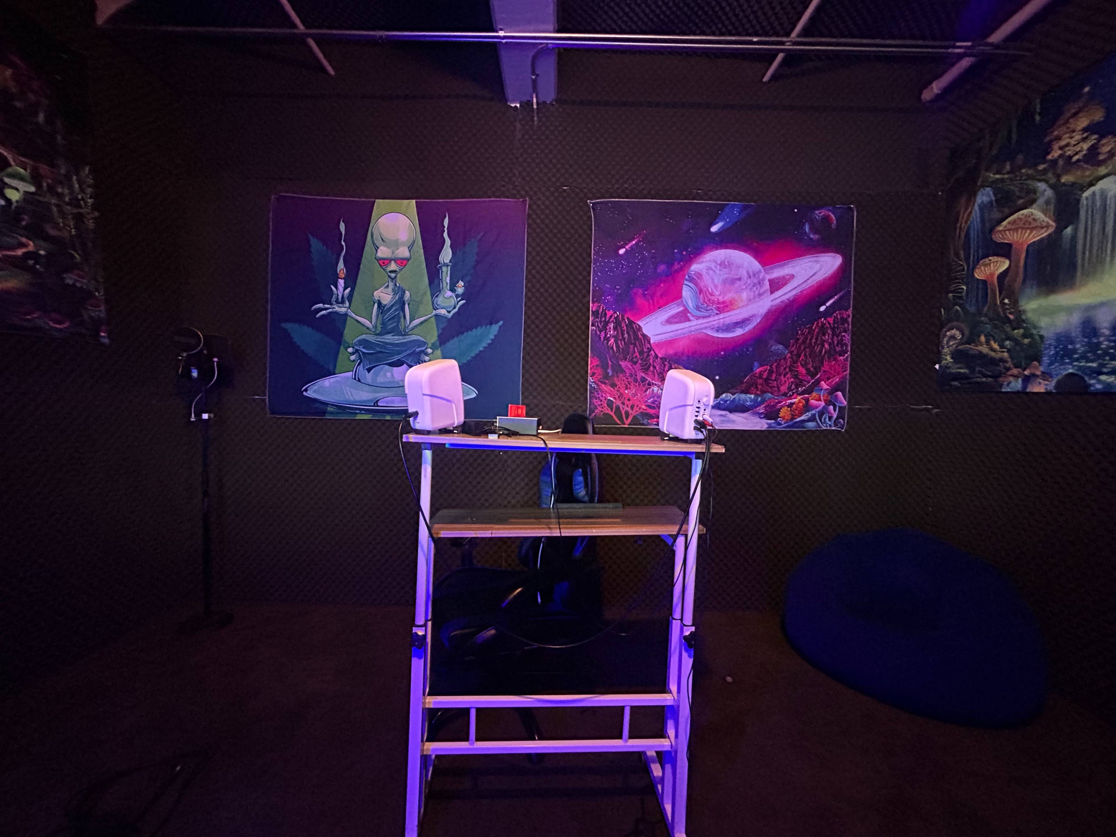 A small room with colorful wall tapestries, a folding table with speakers, and a bean bag chair in the corner.