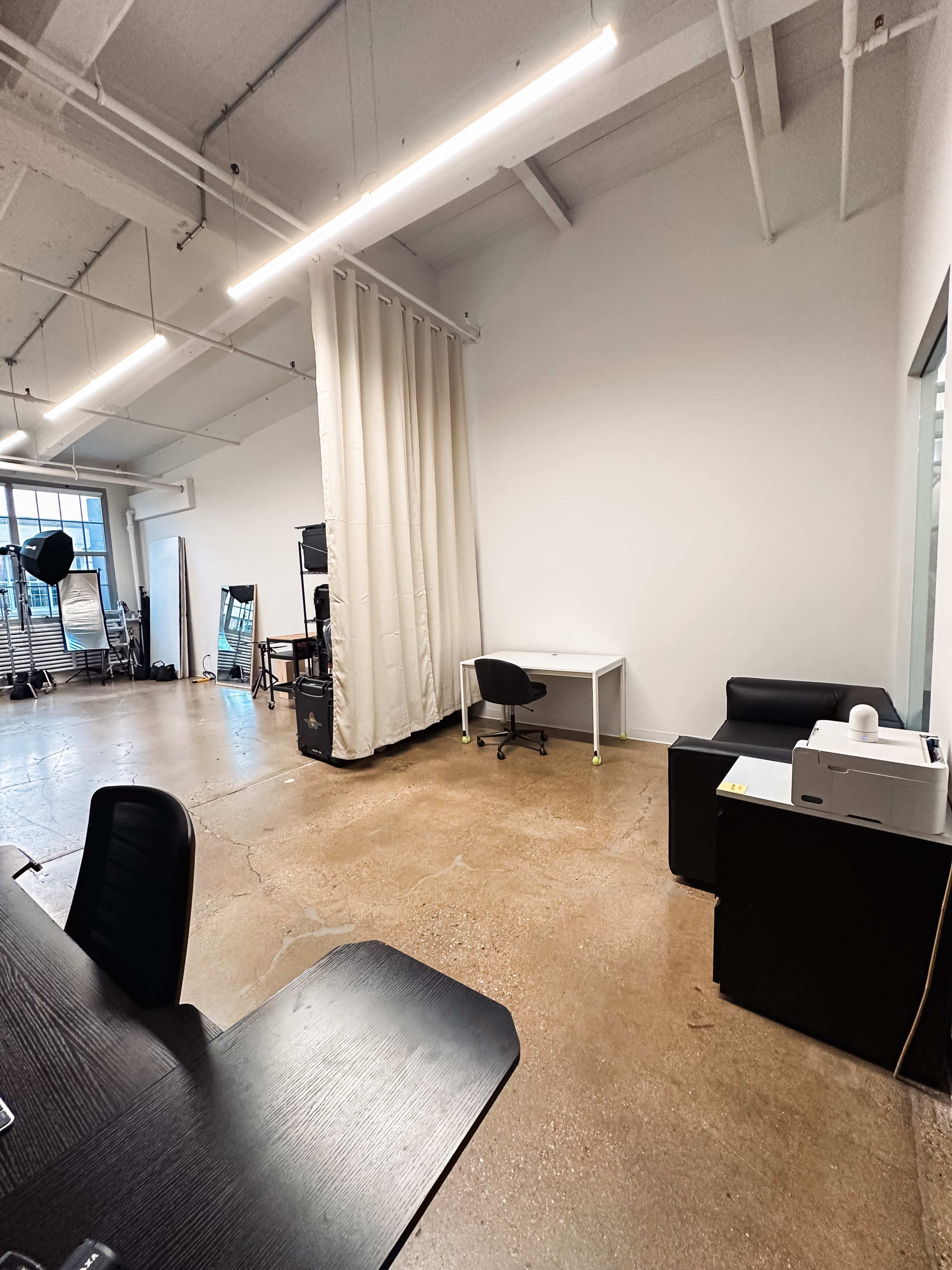 A spacious, modern office with a large window and a desk on one side, while a curtain separates another area featuring a black sofa and a printer.