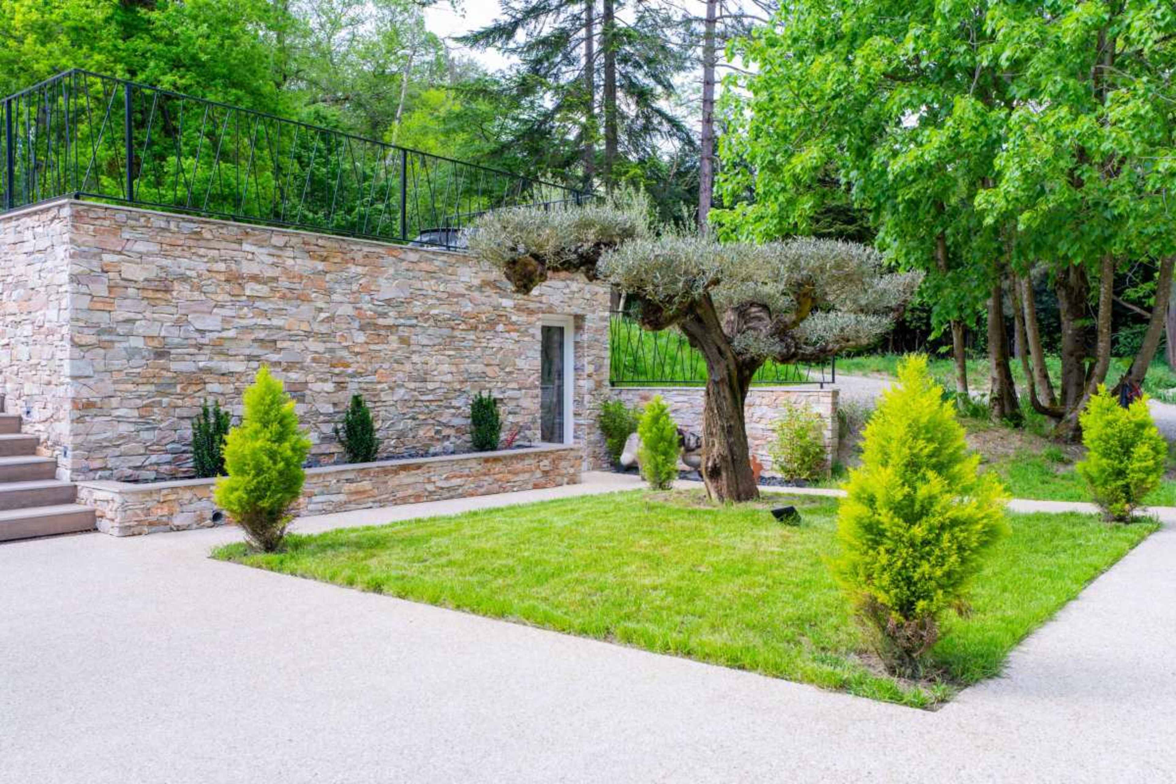 The image shows a landscaped outdoor area featuring a stone wall, manicured shrubs, a grassy lawn, and a decorative tree surrounded by a pathway.