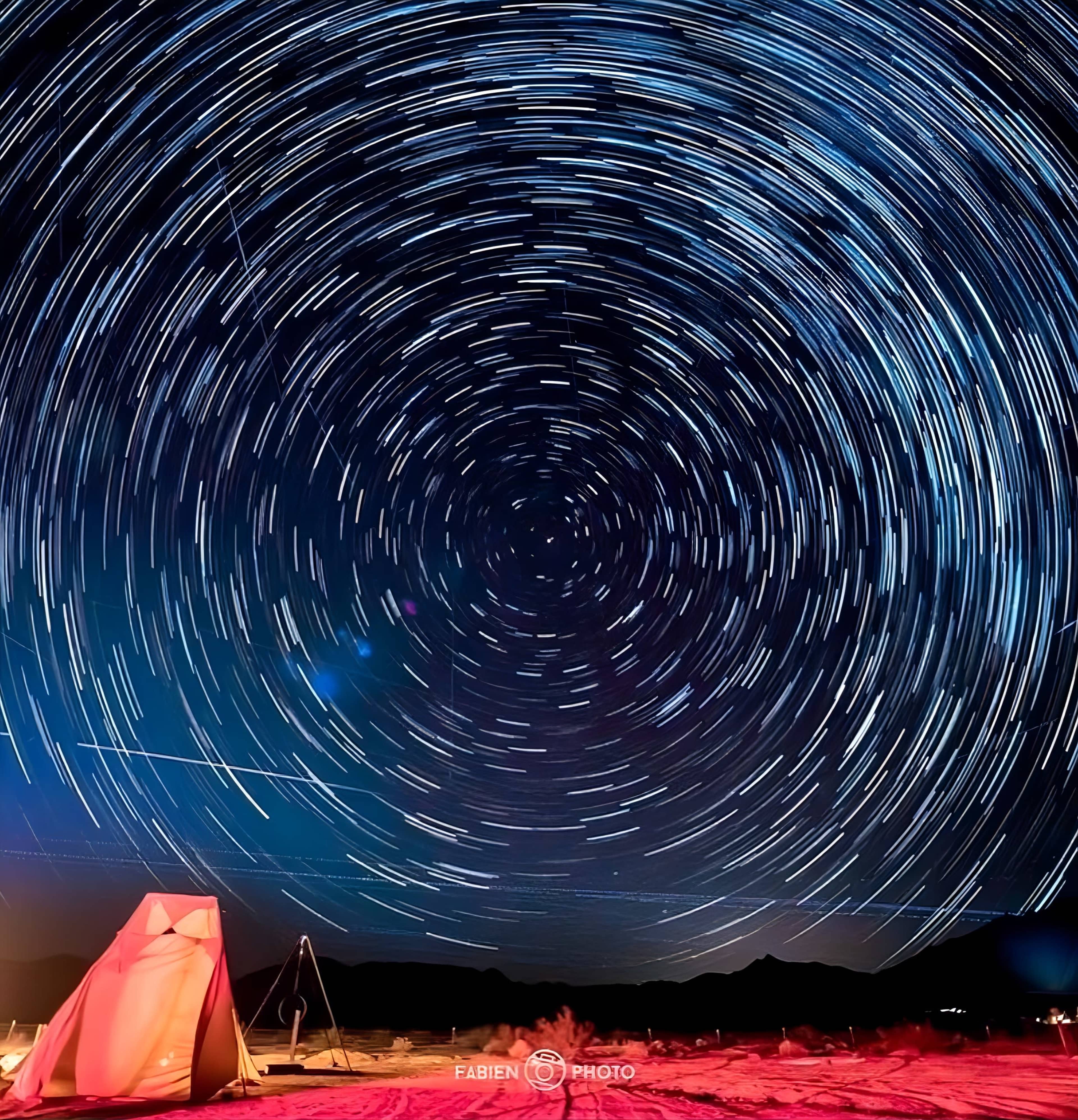 A tent is set up on a sandy landscape under a starry sky, with trails of stars creating a circular motion around a central point.