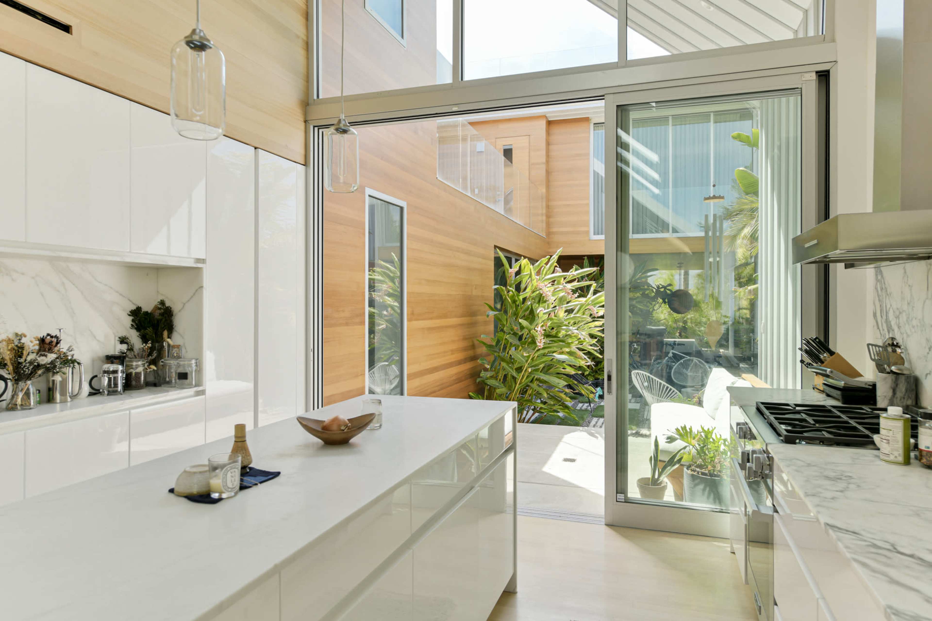A modern kitchen with white cabinetry and a large island, overlooking a bright, green indoor garden through glass sliding doors.