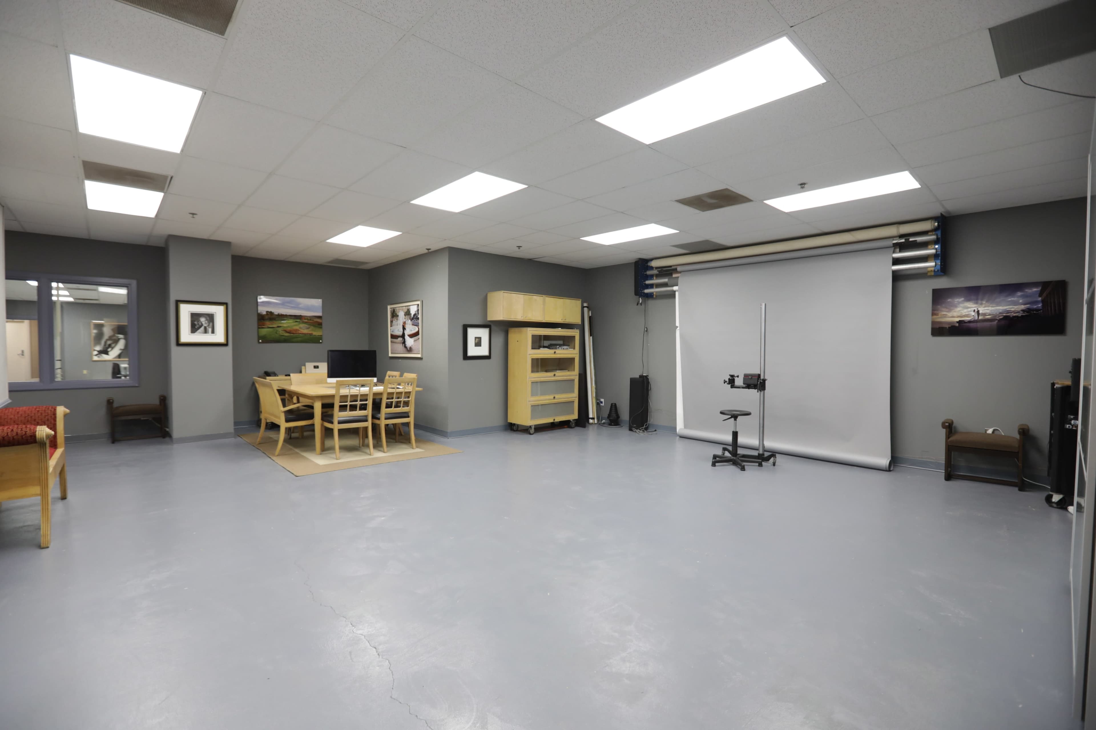 An empty studio space featuring a table with chairs, a shelf, and a backdrop setup against a gray wall.