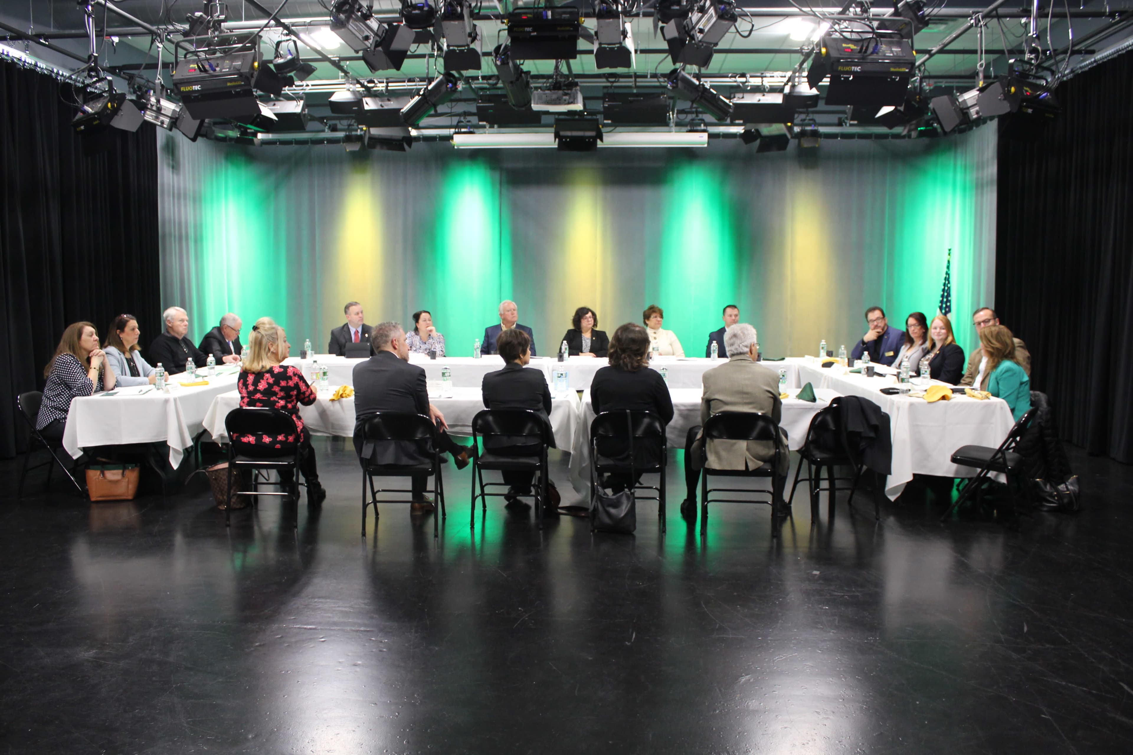 A group of people is seated around a long table in a studio, discussing in a space with green and yellow lighting.