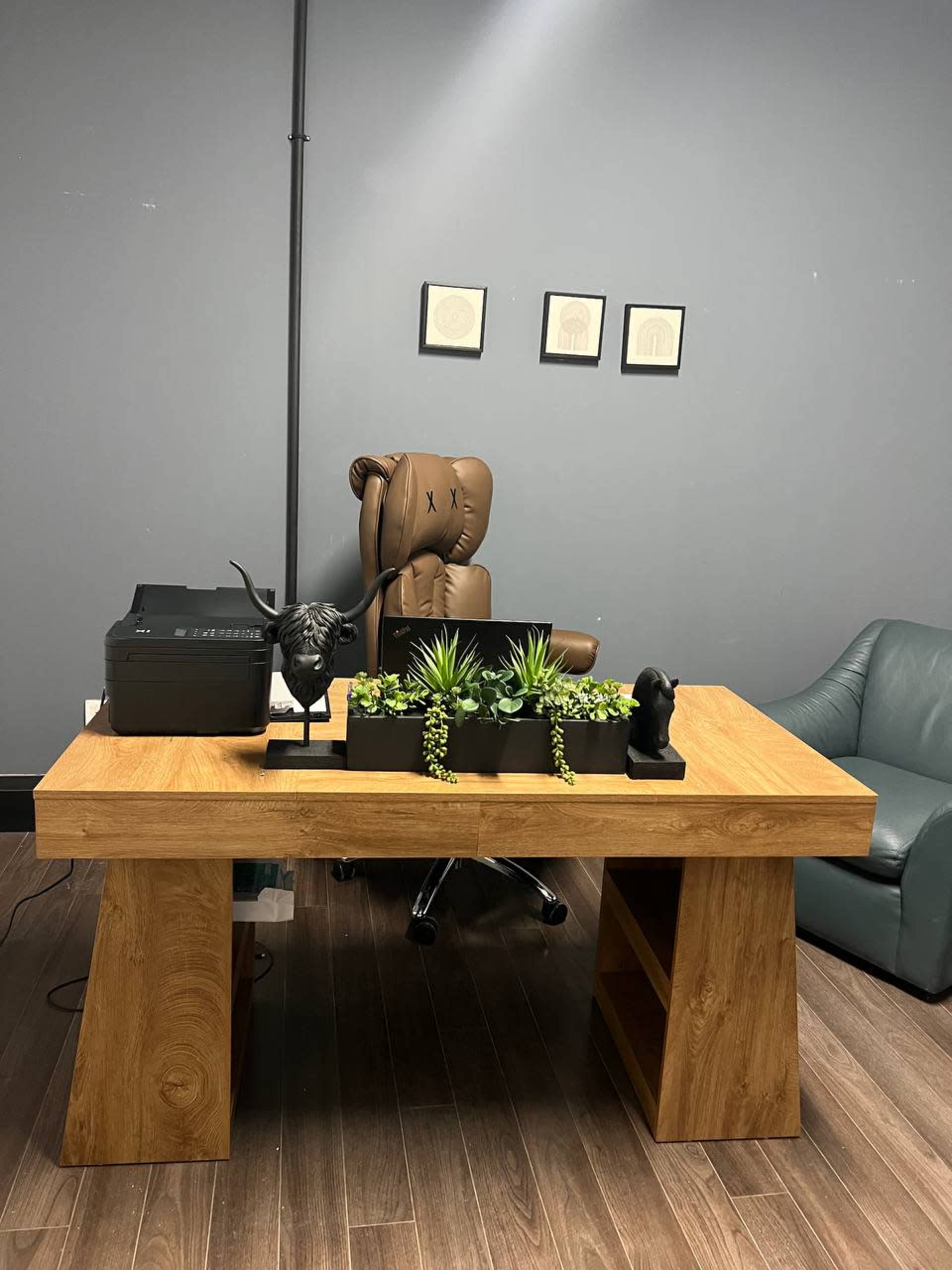 The image shows a modern wooden desk with a printer, small potted plants, decorative figures, and a plush chair in a gray-walled office space.