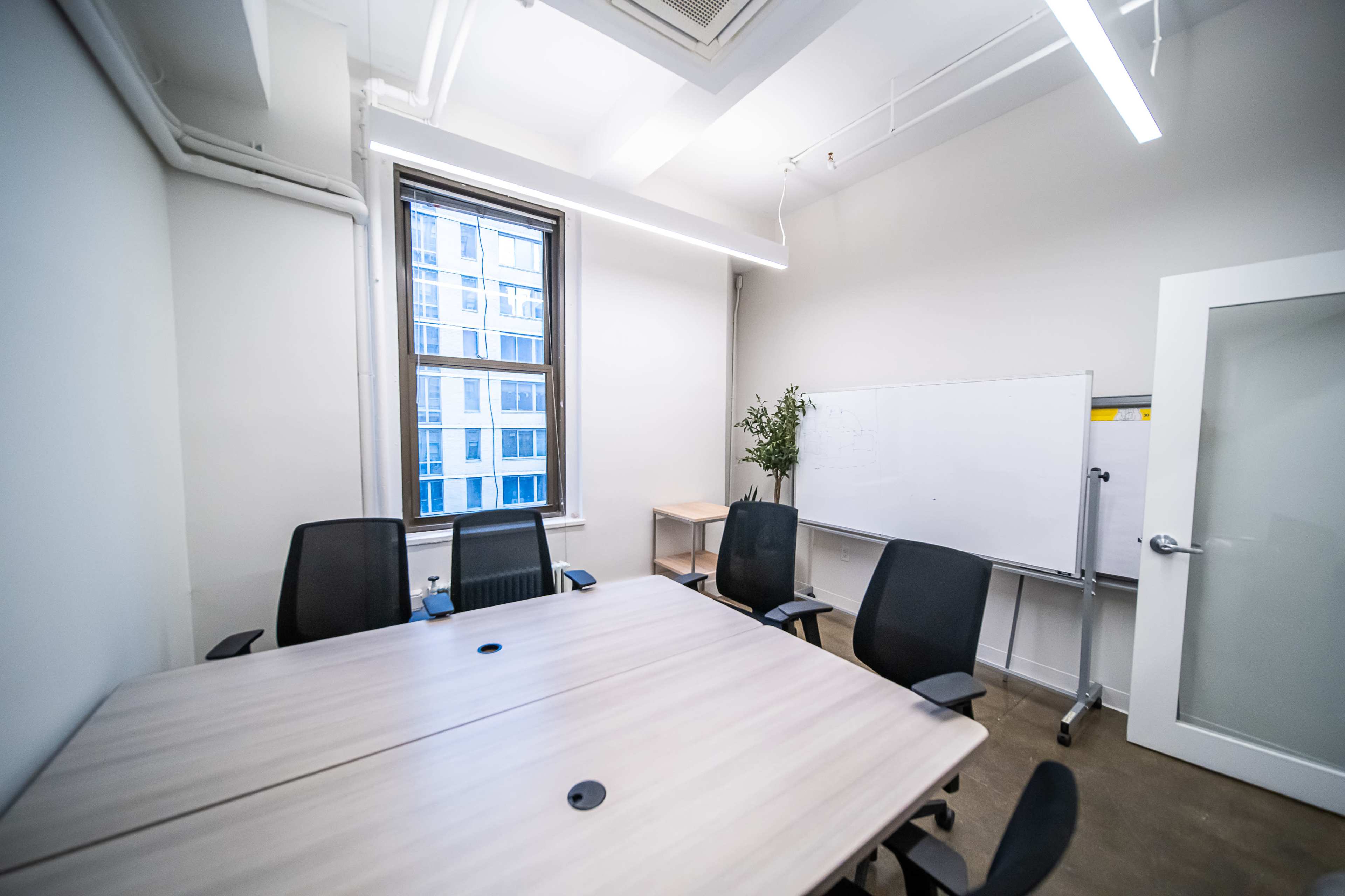 A conference room with a large wooden table, six black chairs, a whiteboard on wheels, and a window with a view of a building outside.
