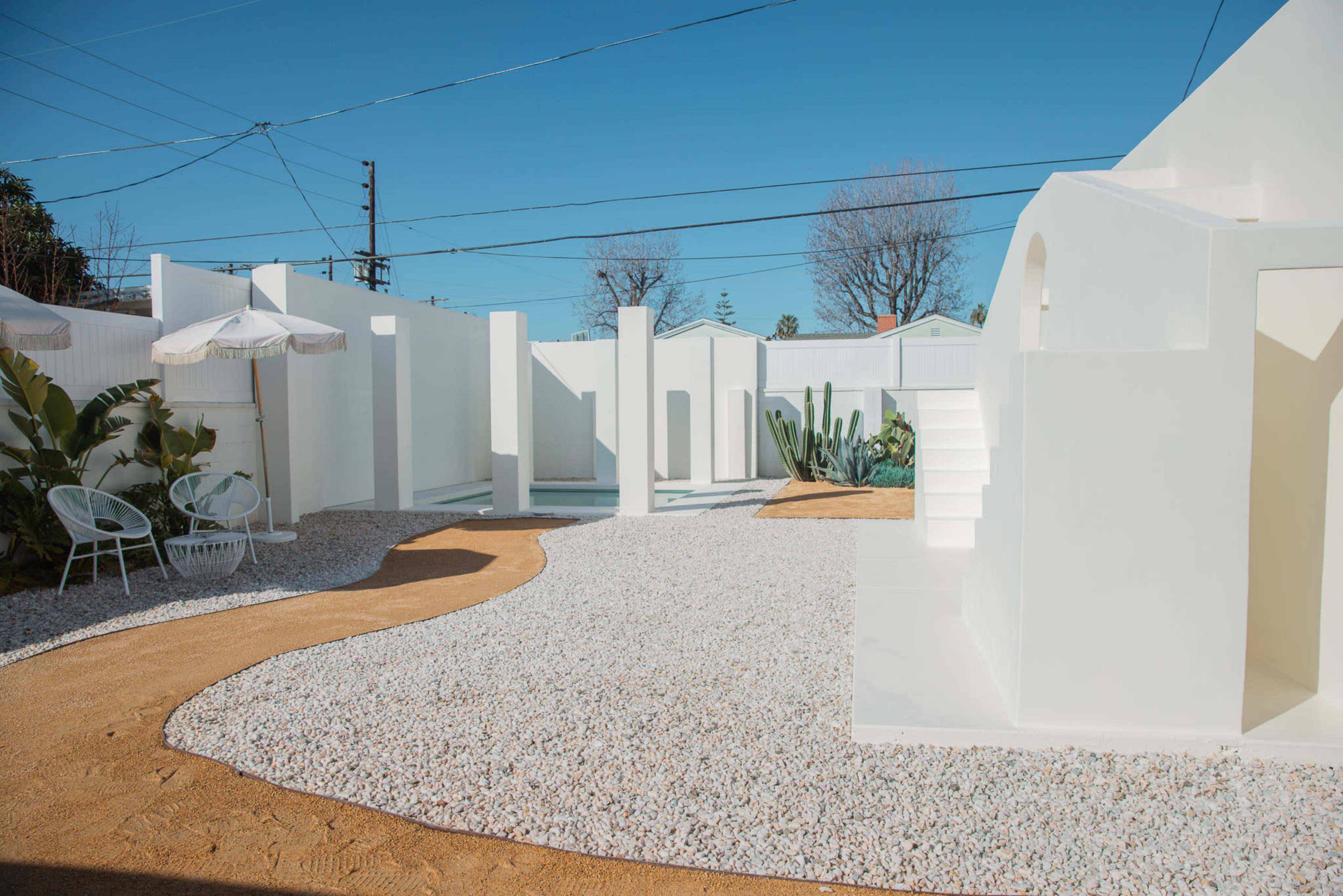 A minimalist outdoor area featuring white walls, gravel pathways, and a small pool surrounded by greenery and seating.