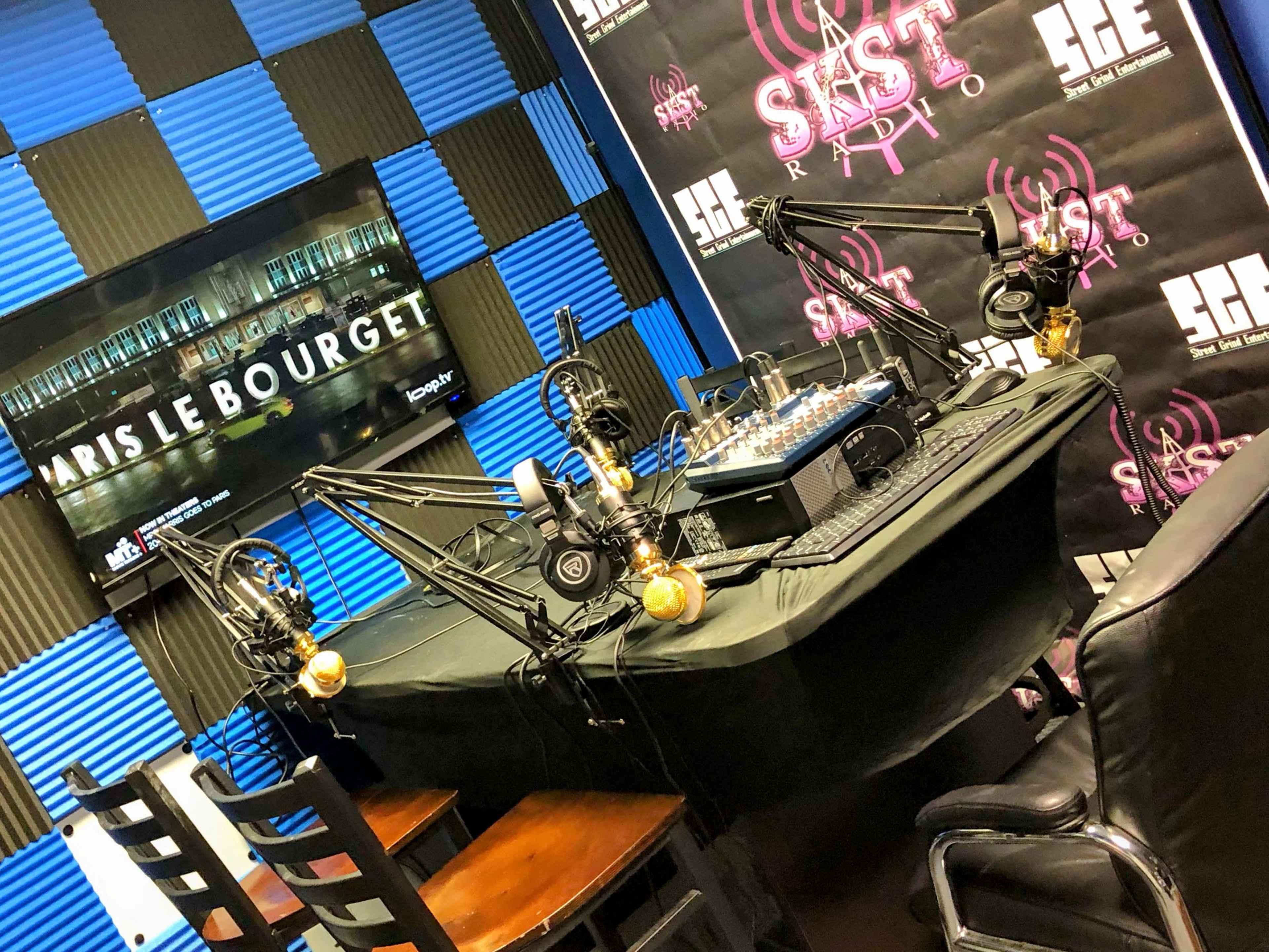 A radio studio is set up with multiple microphones, a soundboard, and a large screen displaying a logo against a blue and black patterned background.