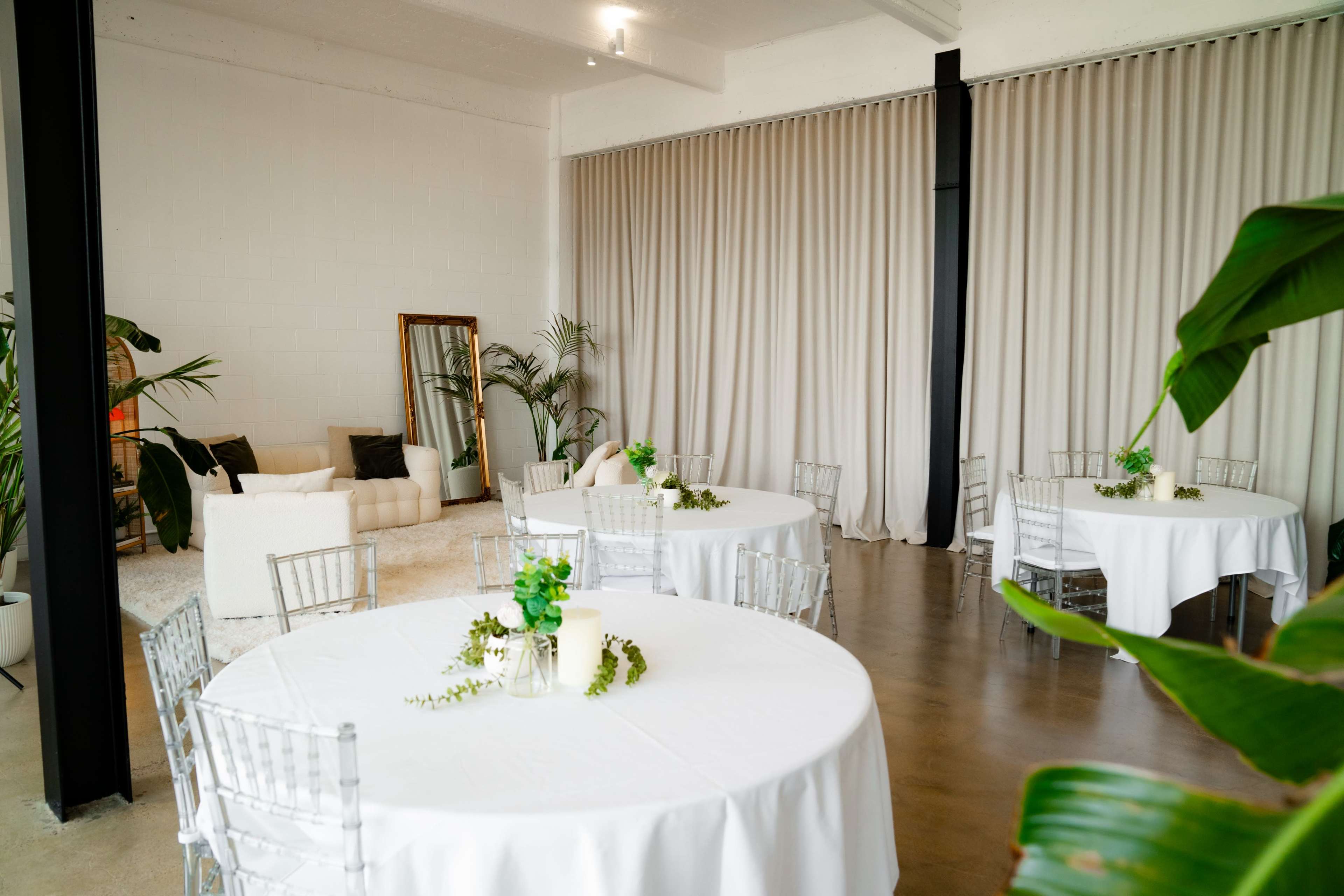 The image shows a modern event space with round tables covered in white tablecloths, a sofa, and plants, along with a large mirror and curtains.
