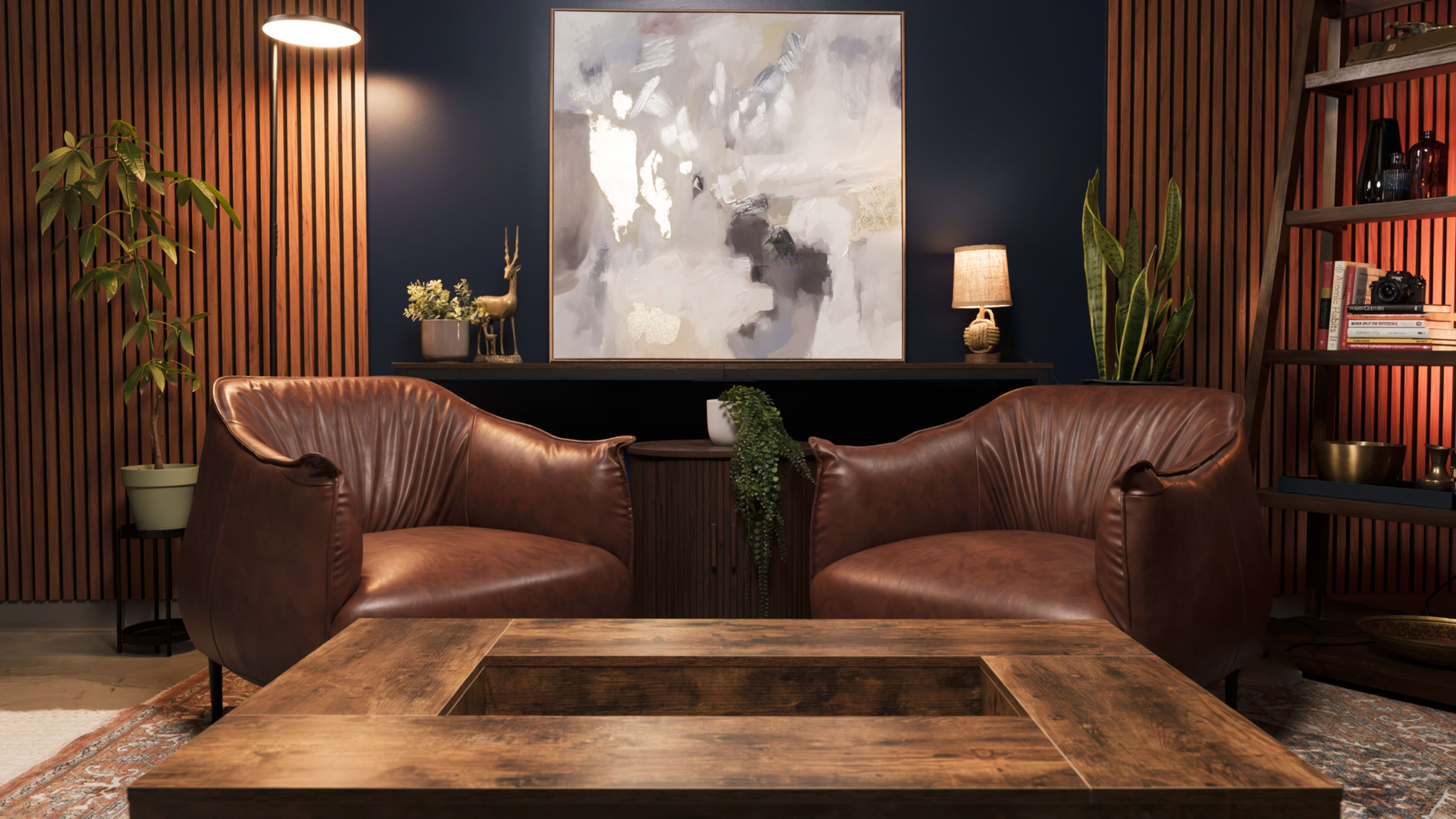 The image features a modern living room with two brown leather chairs facing a wooden coffee table, a large abstract painting on the dark blue wall, and various decorative elements, including a lamp, plants, and a shelf.