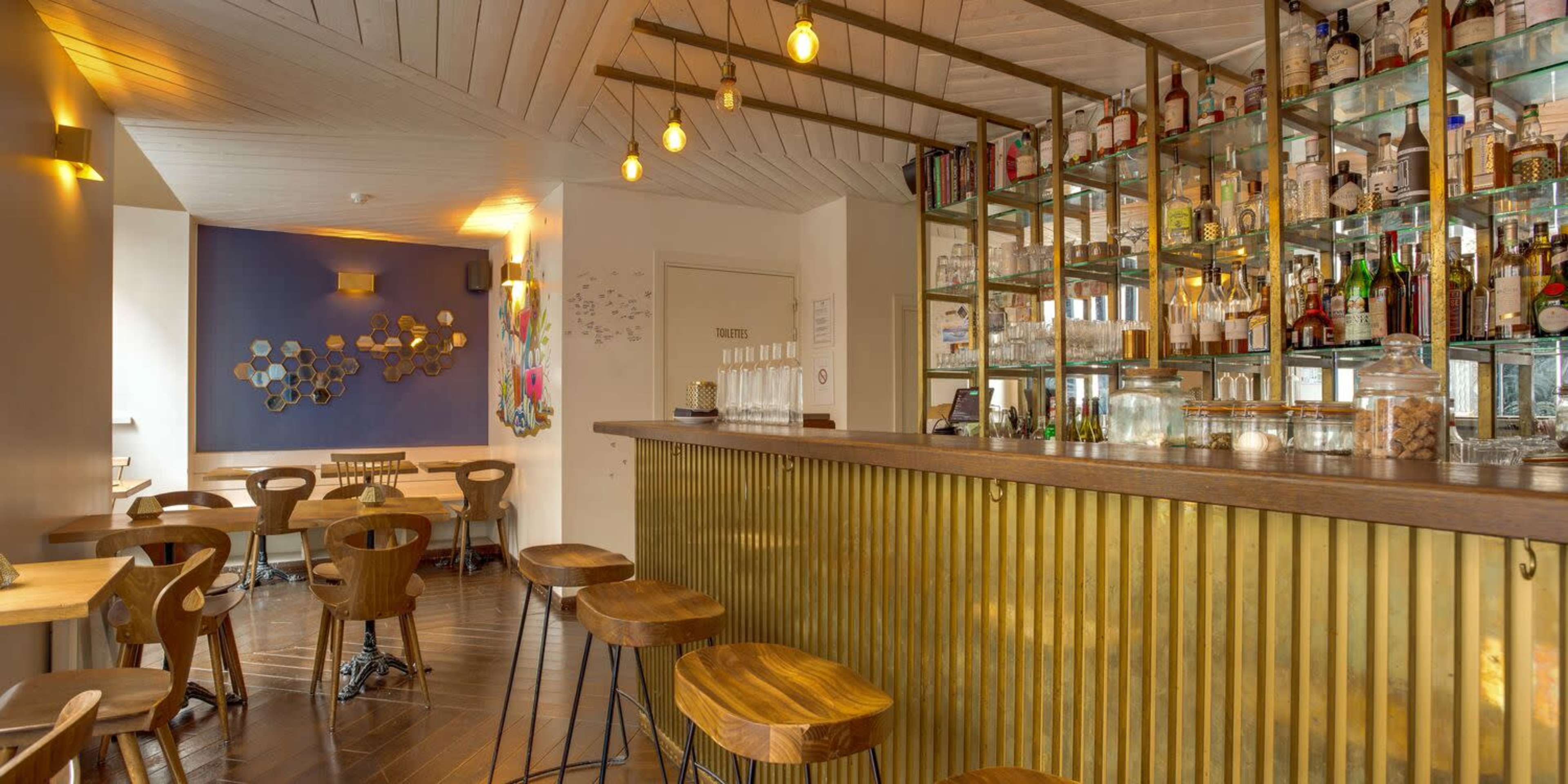 A modern bar interior with a wooden counter, high stools, shelves filled with various bottles, and a few tables scattered throughout the space.