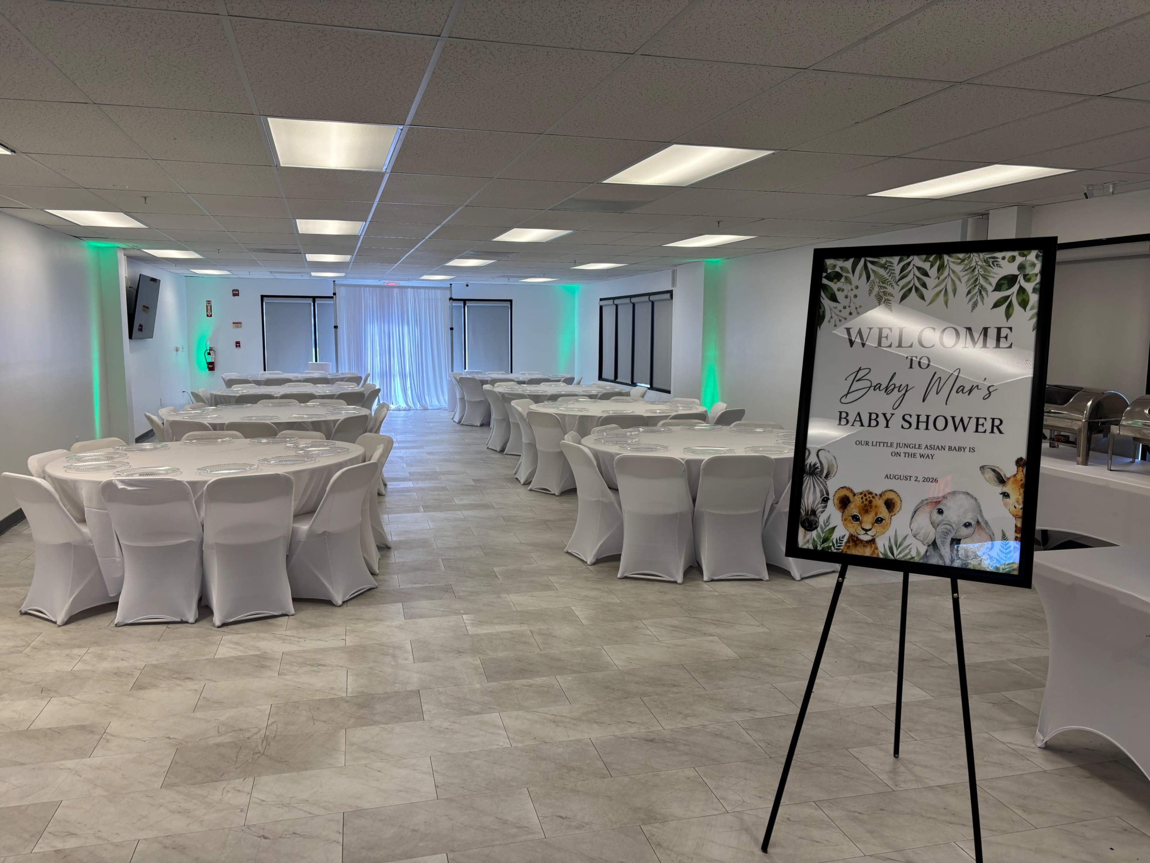 The image shows a spacious banquet hall set up for a baby shower, with round tables covered in white tablecloths and a welcome sign featuring animal illustrations.