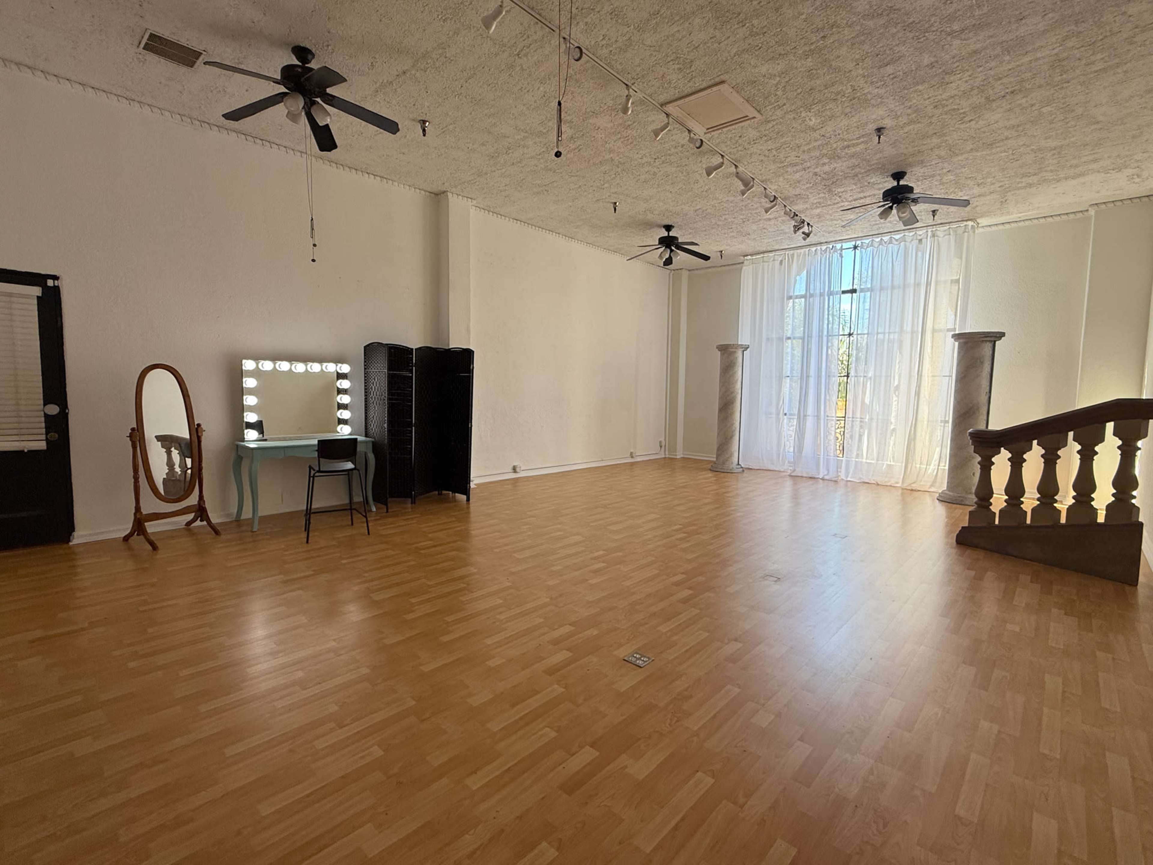 The image shows a spacious, well-lit interior room featuring a large window with sheer curtains, a mirror with lights, a chair, and a staircase leading to a balcony.