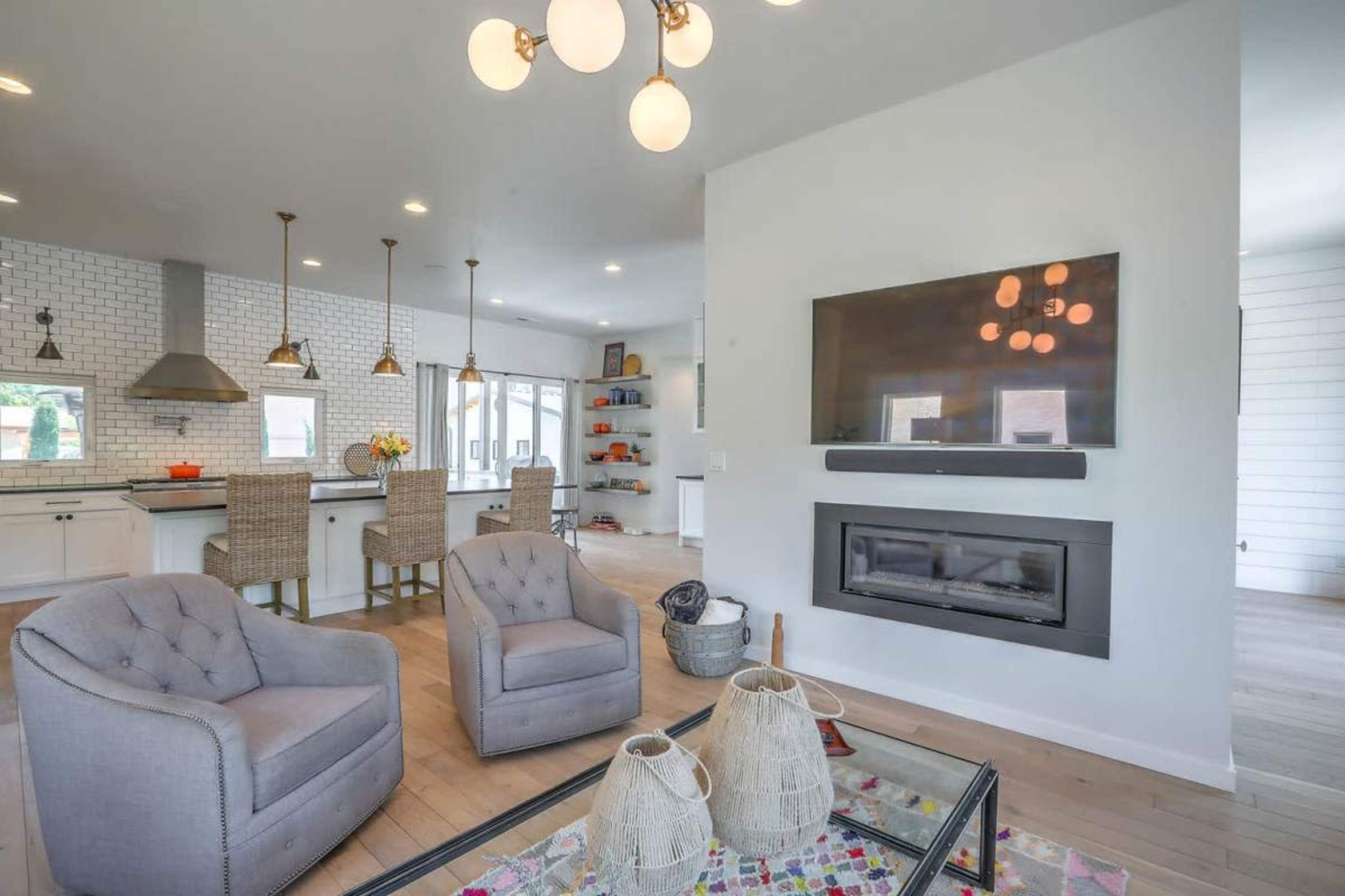 A modern open-concept living area features a fireplace, two upholstered chairs, and a kitchen with bar seating.