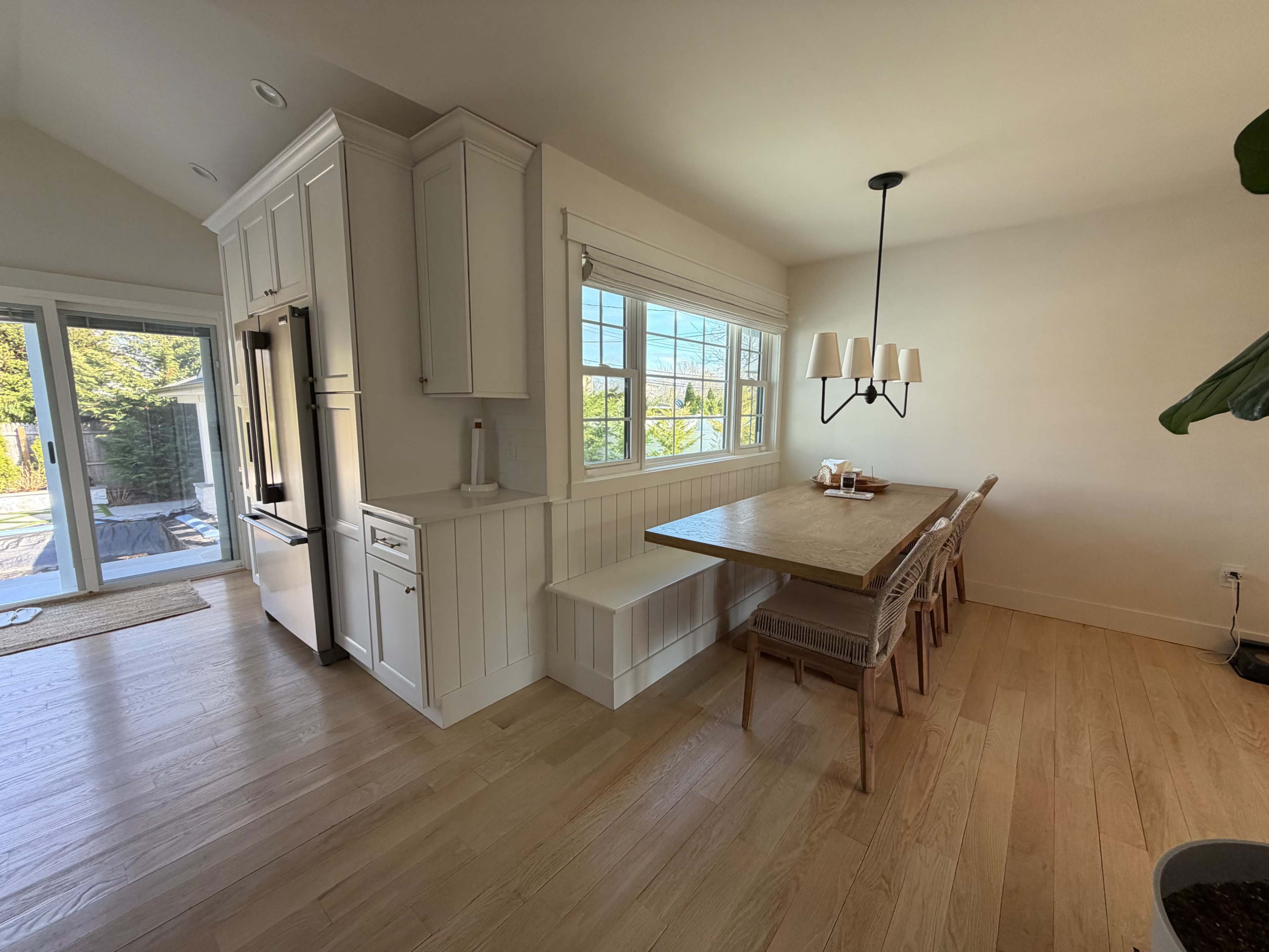 The image shows a modern kitchen and dining area with light wood flooring, a large dining table, and white cabinetry featuring a window with natural light.