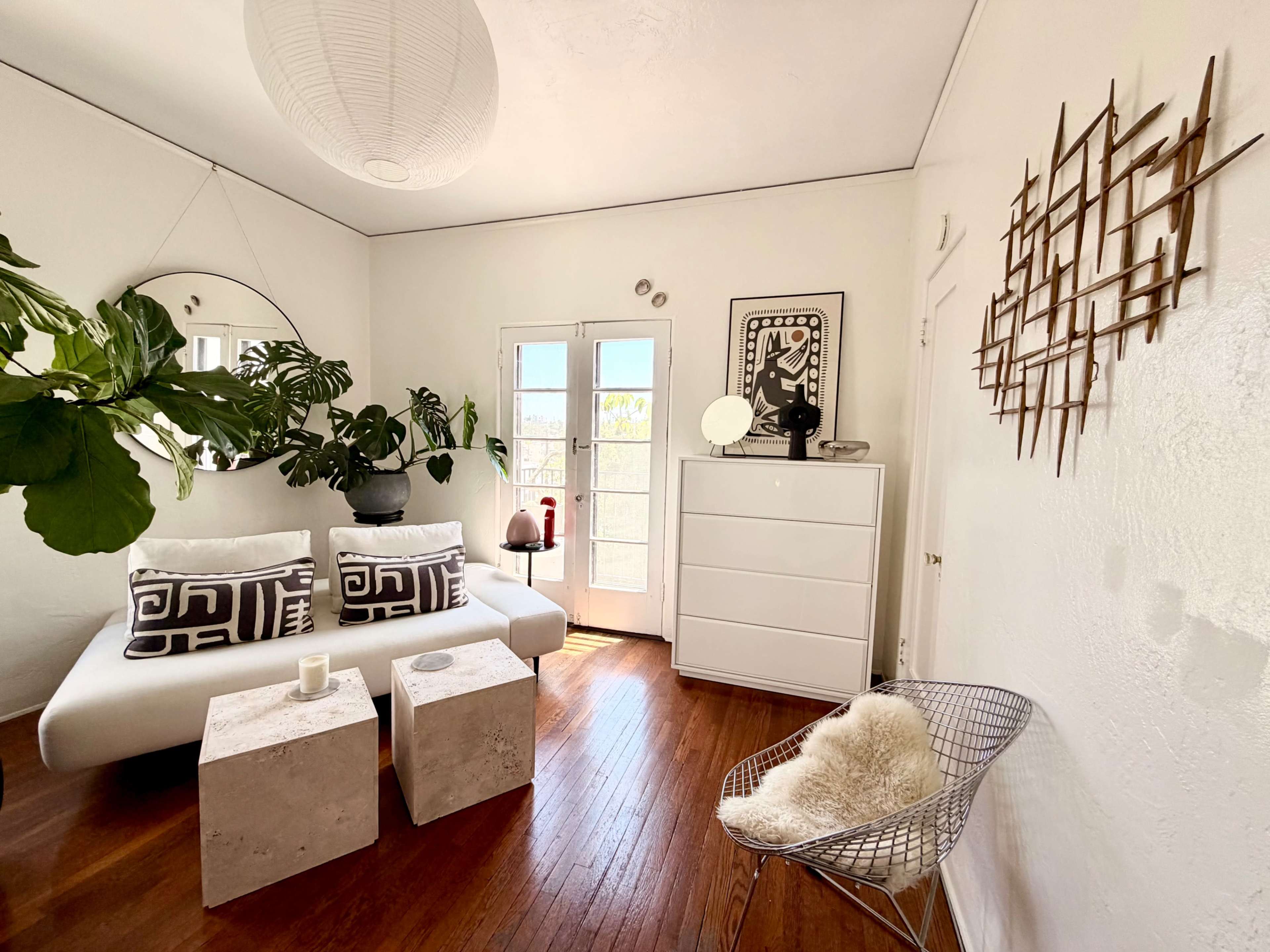 The image shows a bright living room featuring a white sofa, two concrete side tables, a large round mirror, and a potted plant, with light streaming in from French doors.