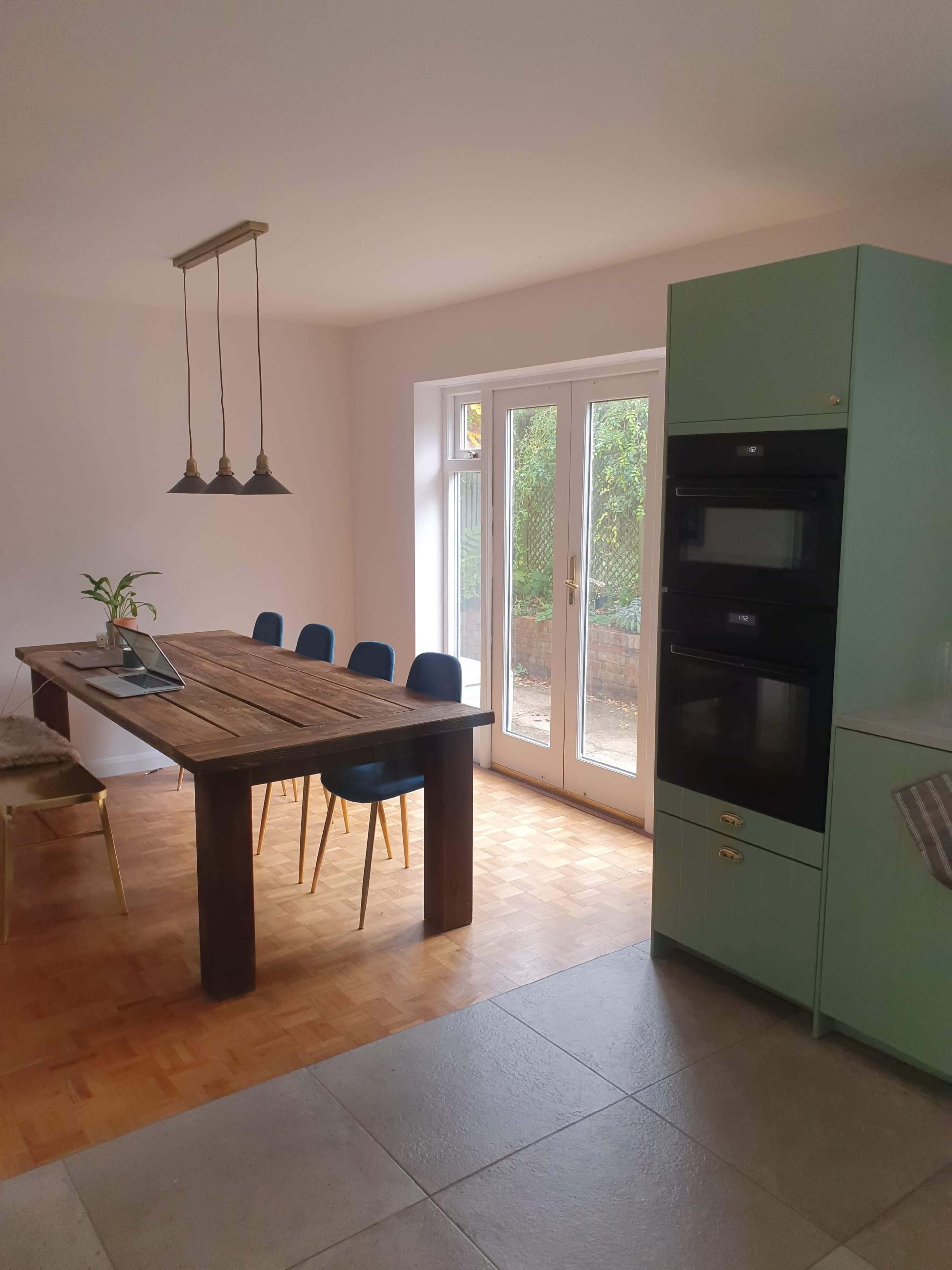 A modern kitchen with a wooden dining table, four blue chairs, and two built-in ovens adjacent to a sliding glass door.