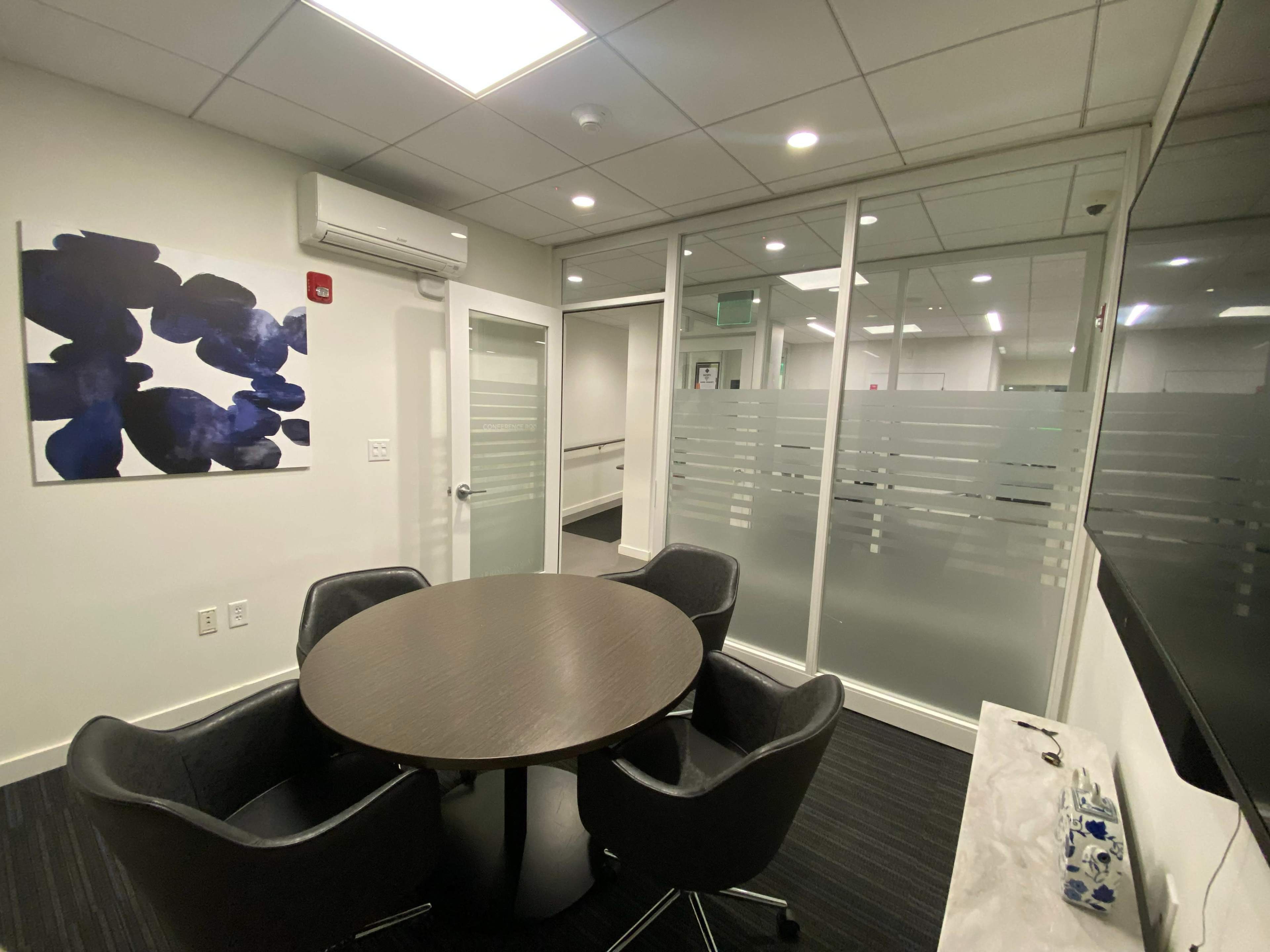 The image shows a modern office meeting room with a round table and four chairs, separated by glass walls from the adjacent area.