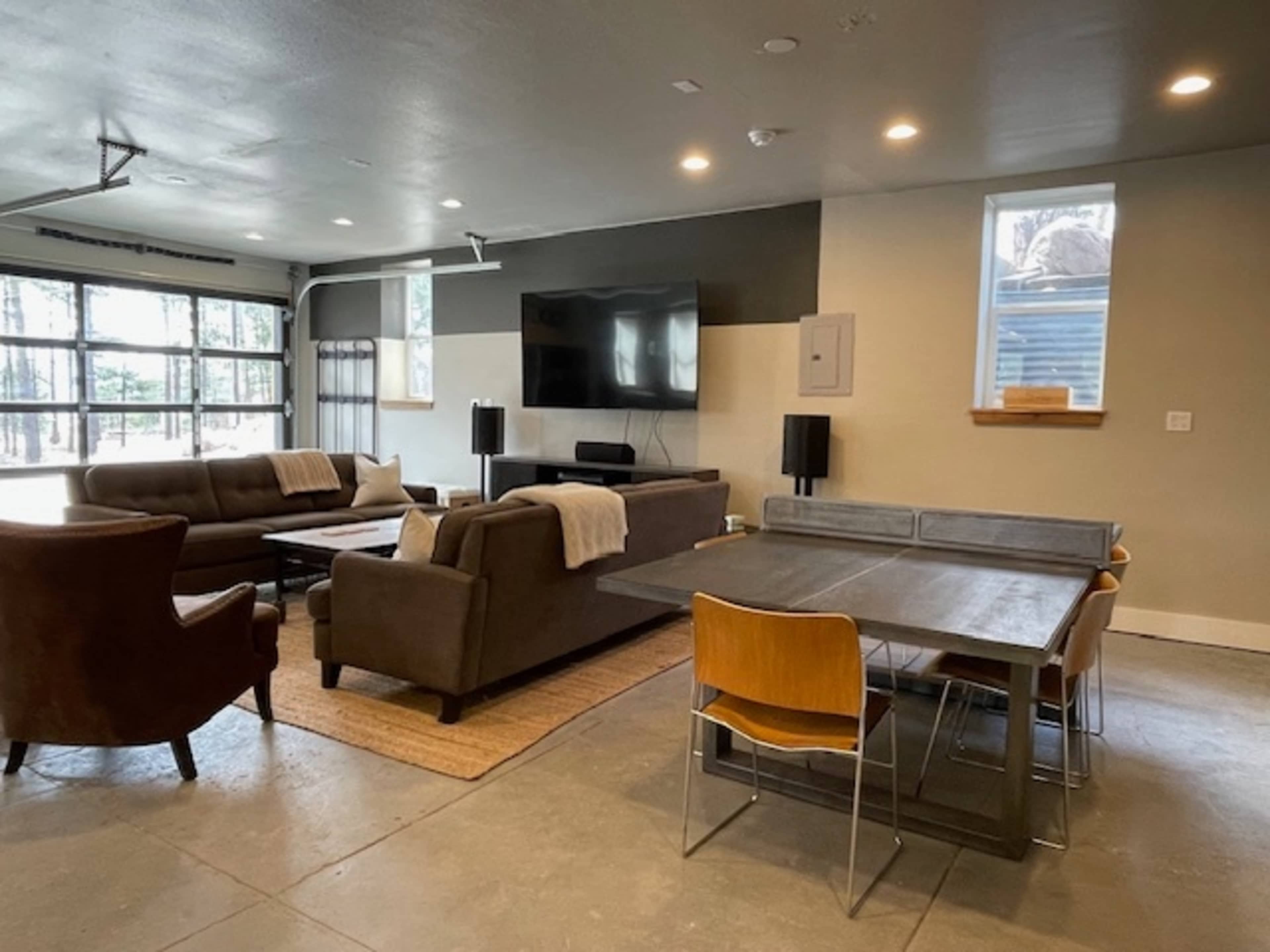 A modern living space features a sectional sofa, armchair, large TV, and dining table with chairs against a backdrop of large windows and minimalistic decor.