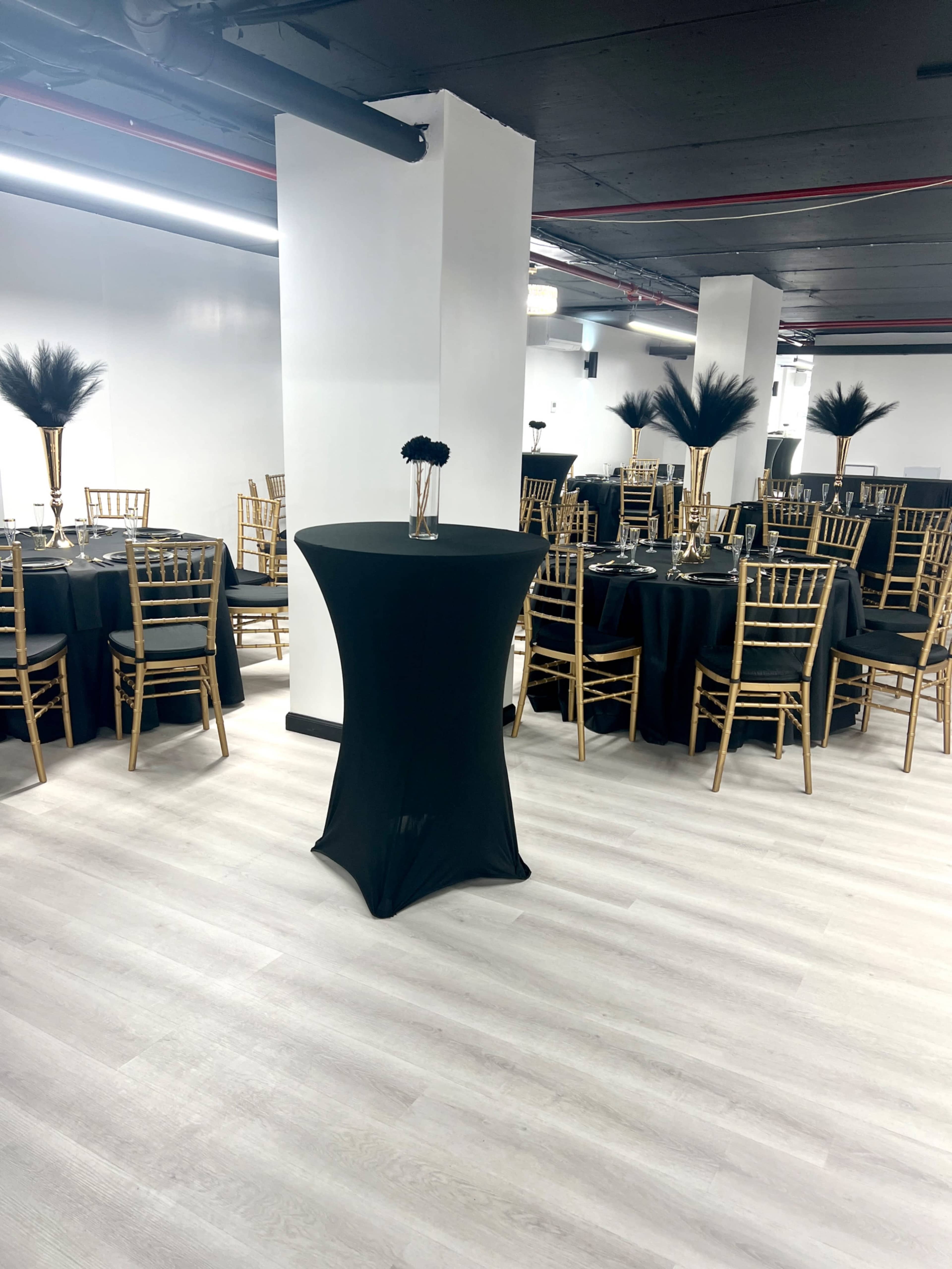 A spacious event room set with round tables covered in black tablecloths, each adorned with gold chairs and centerpieces featuring tall vases.